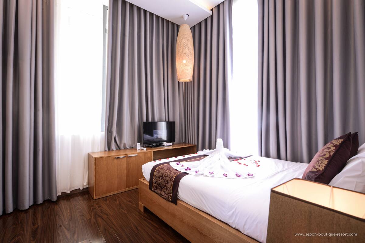 Ein Hotelzimmer mit einem hölzernen Bett, einem Fernseher auf einem Nachttisch aus Holz und grauen Vorhängen.