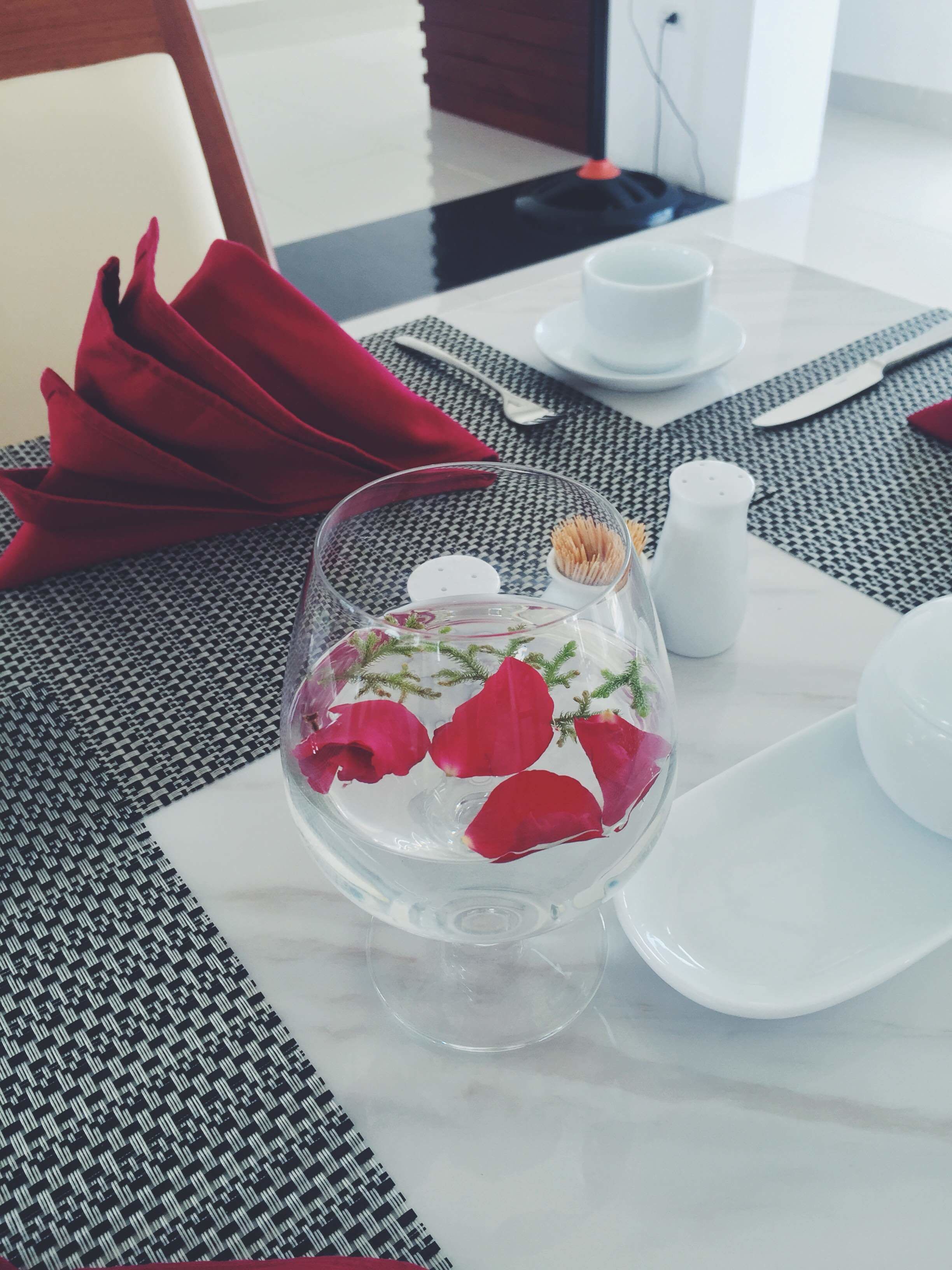 A table with a clear glass vase containing red rose petals, a white salt shaker, and a white cup.