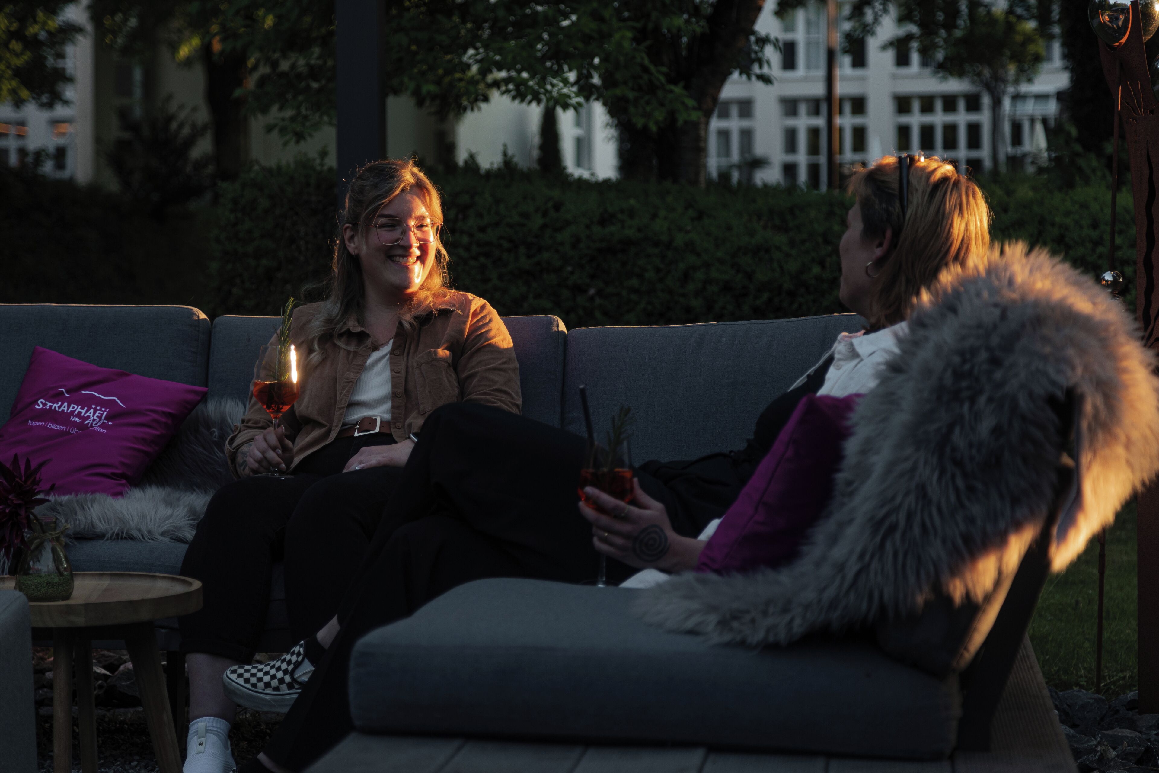 Zwei Personen sitzen auf einem grauen Sofa mit einem violetten Kissen und halten Getränke.