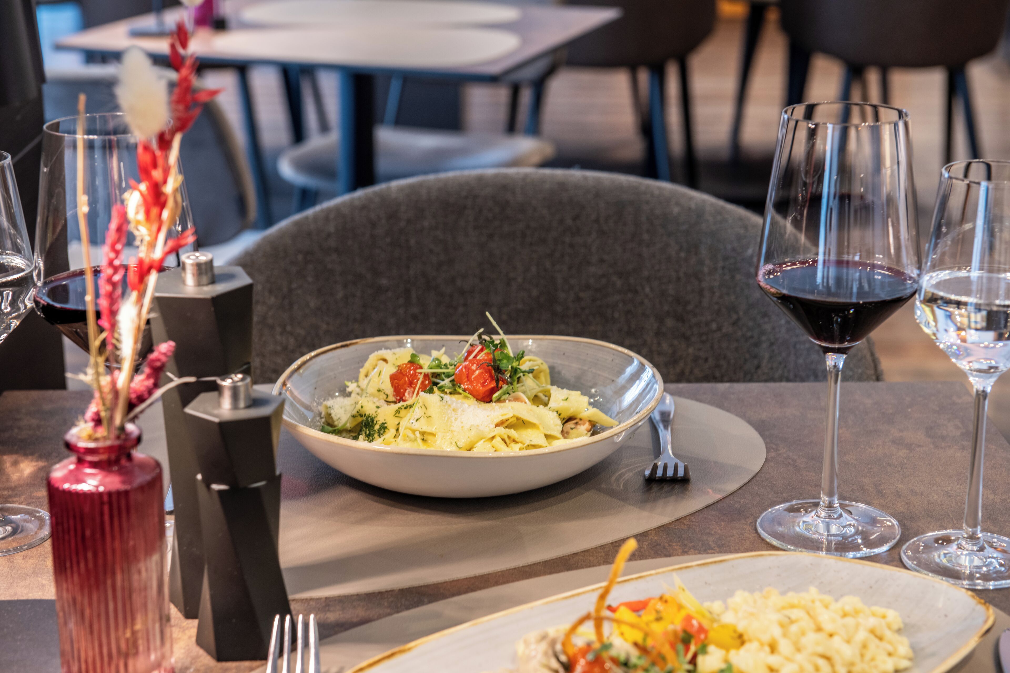 Ein Esstisch mit einer Pasta-Schüssel, zwei Weingläsern und einer roten Vase mit Blumen.