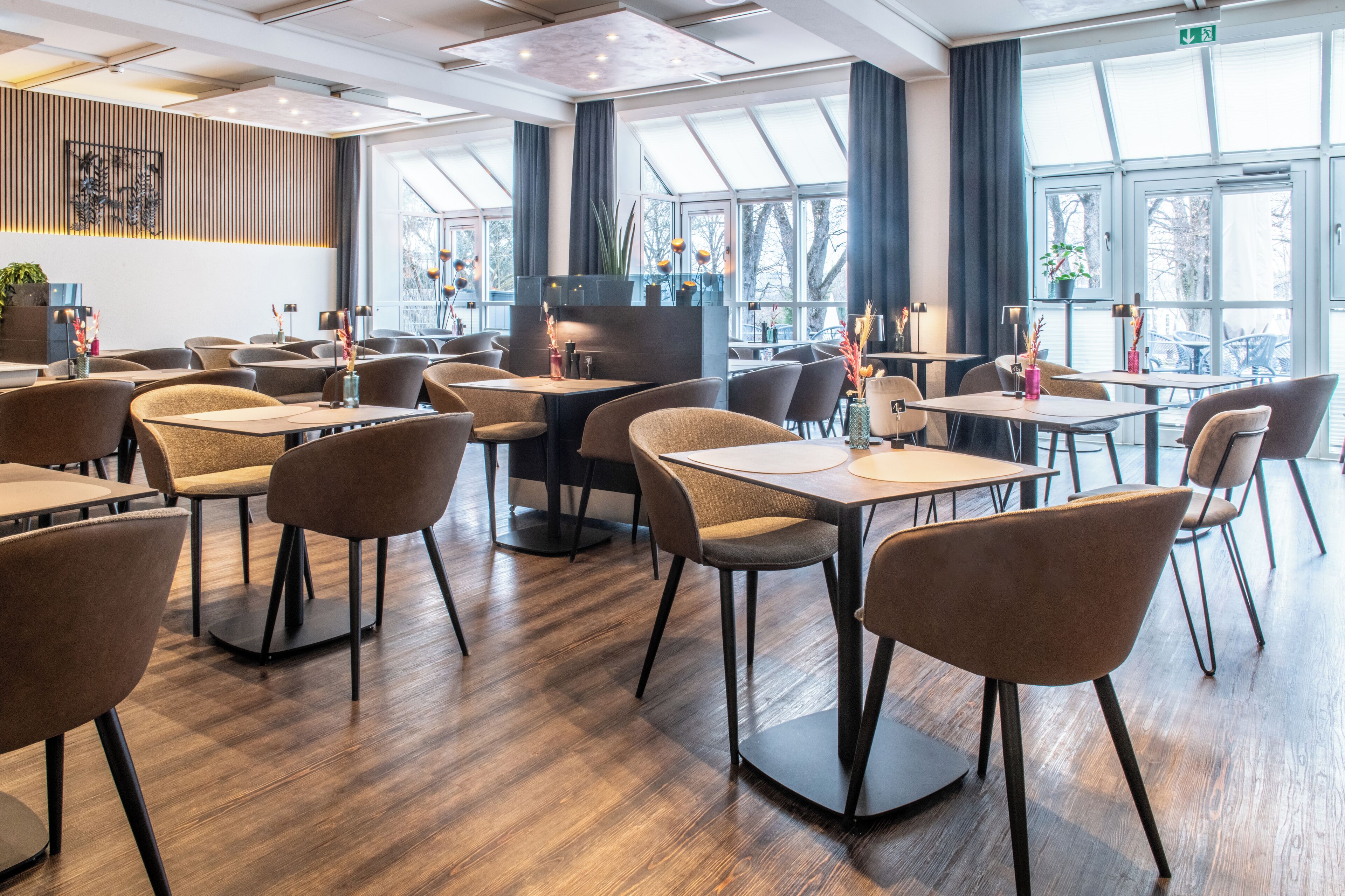 A modern dining area with multiple tables and chairs, large windows with curtains, and a wooden floor.