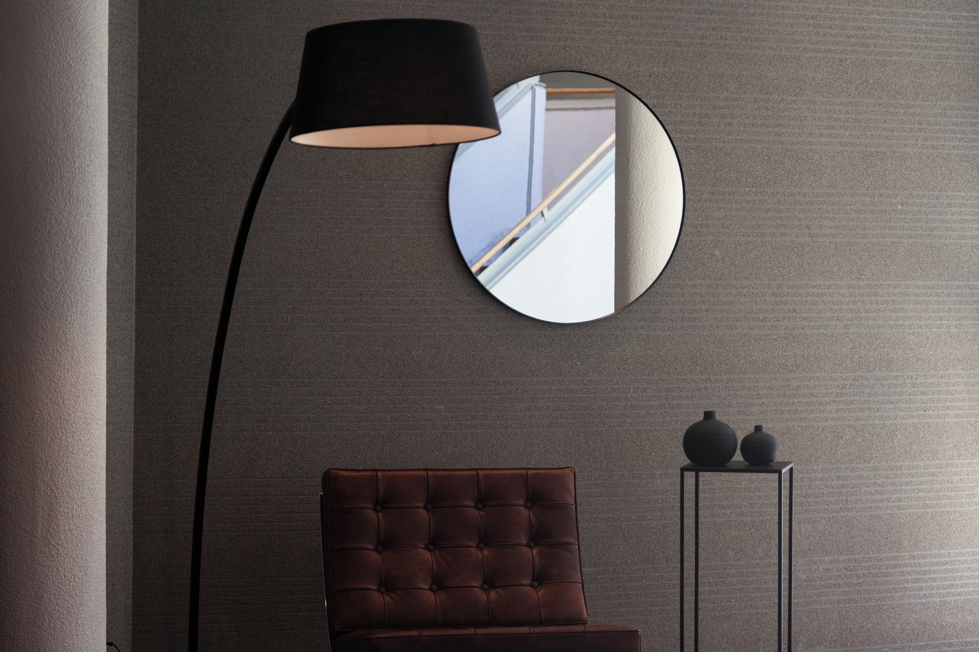 Una habitación con una butaca color café con textura, una lámpara de pie negra, un espejo redondo y una mesita con dos jarrones.