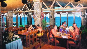 Un restaurante rústico frente al mar, con mobiliario de madera y grandes ventanales con vistas al océano.