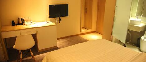 A hotel room with a bed, a desk with a computer, a chair, a television, and a bathroom with a sink and mirror.