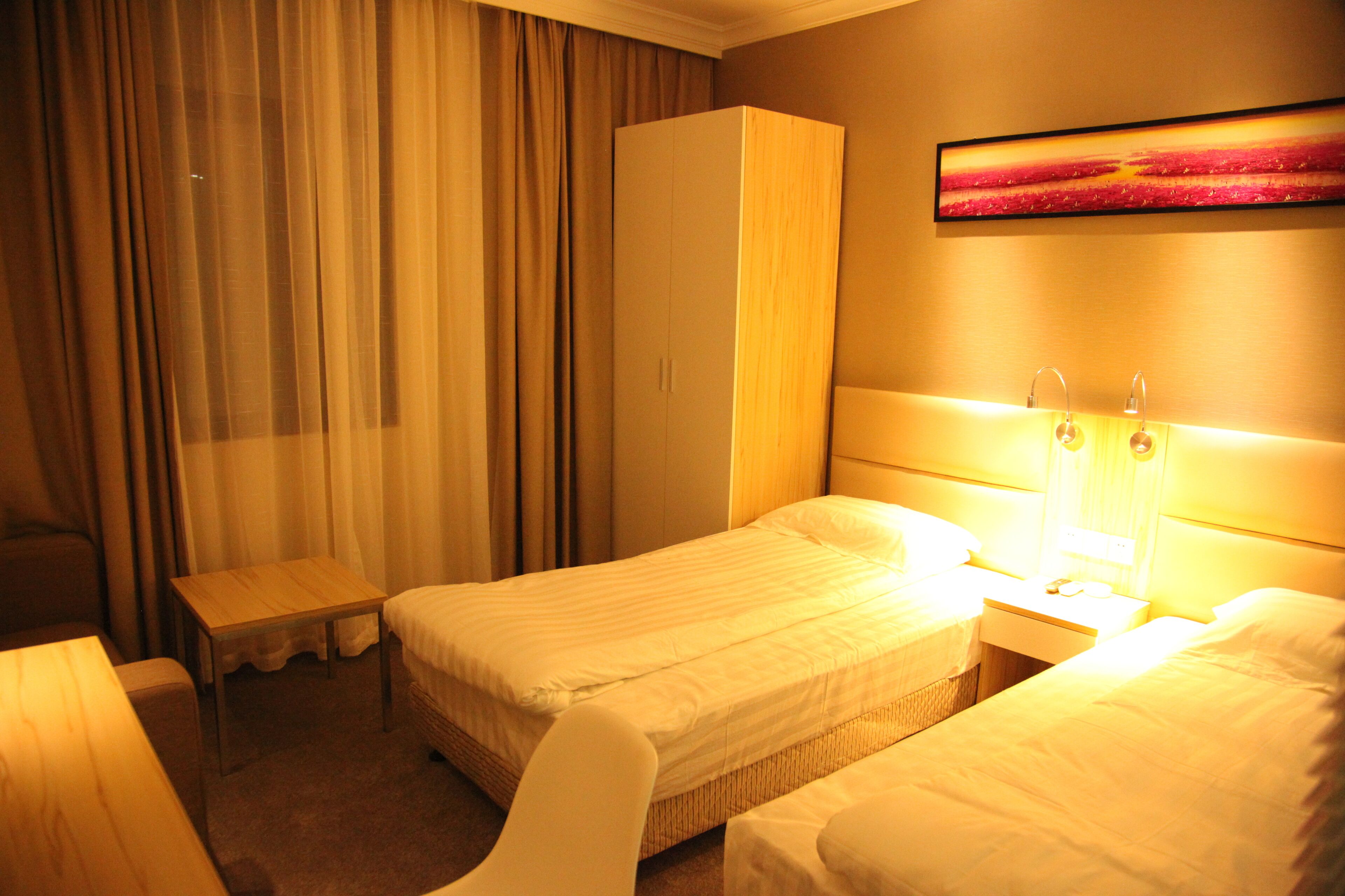 Une chambre d’hôtel avec deux lits simples, une table de chevet en bois, une petite table, une armoire et un tableau au mur.