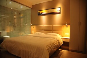 A modern bedroom with a glass shower enclosure, a large bed with a headboard, and a painting above the bed.