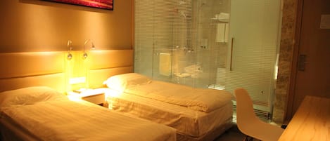 Hotellihuone, jossa on kaksi erillistä sänkyä, yöpöytä lampun kanssa, tuoli ja lasiseinällä erotettu suihkutila.