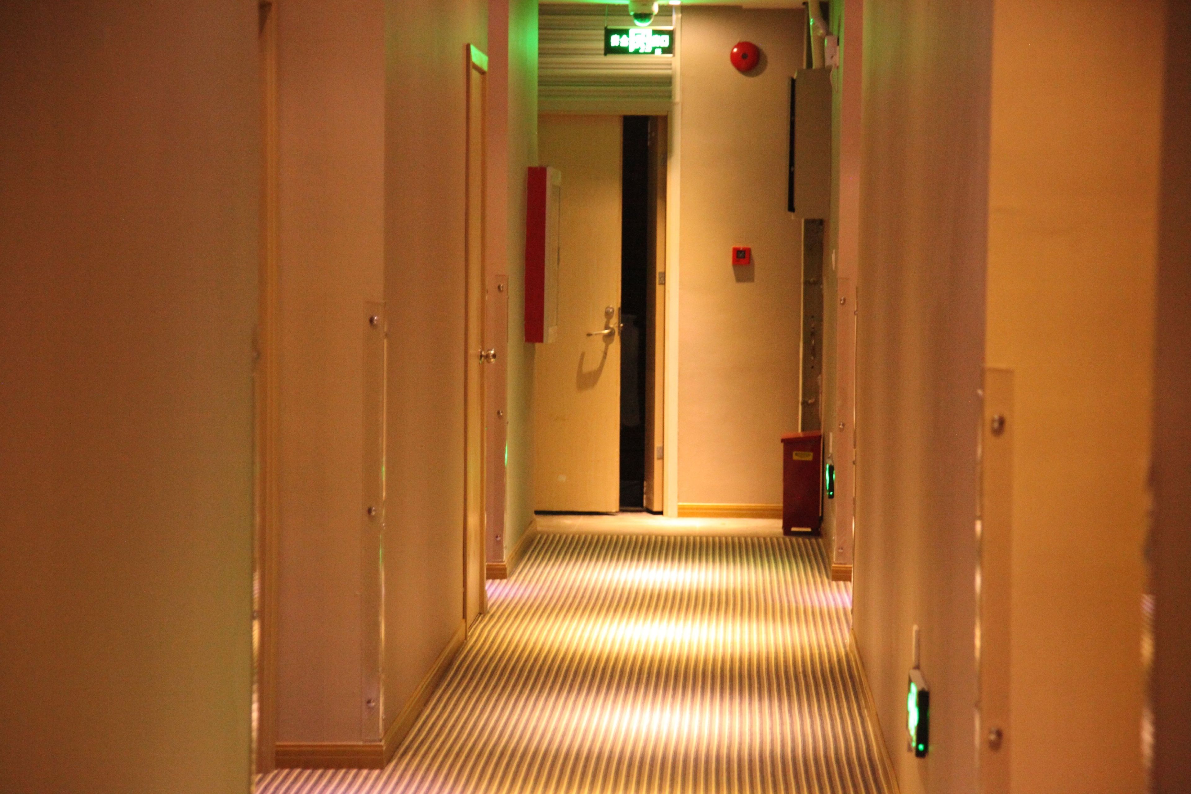 A hotel hallway with a striped carpet, doors on either side, and emergency equipment on the walls.