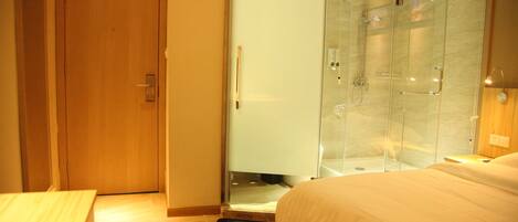 Une chambre d’hôtel avec une porte en bois, une cabine de douche en verre et un lit recouvert d’une couette blanche.
