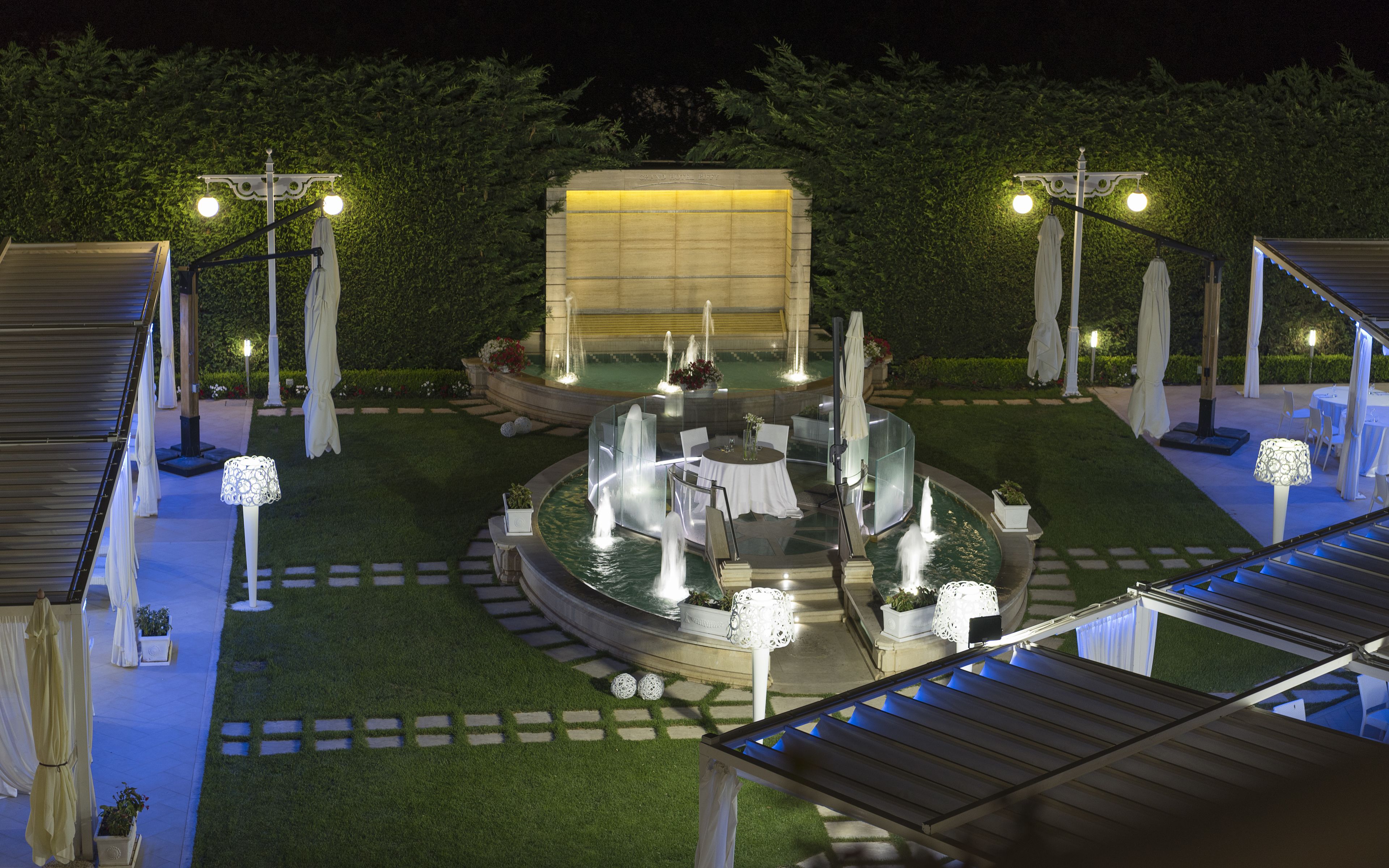 An outdoor patio area with a central water feature, surrounded by tables and chairs, and illuminated by overhead lights.