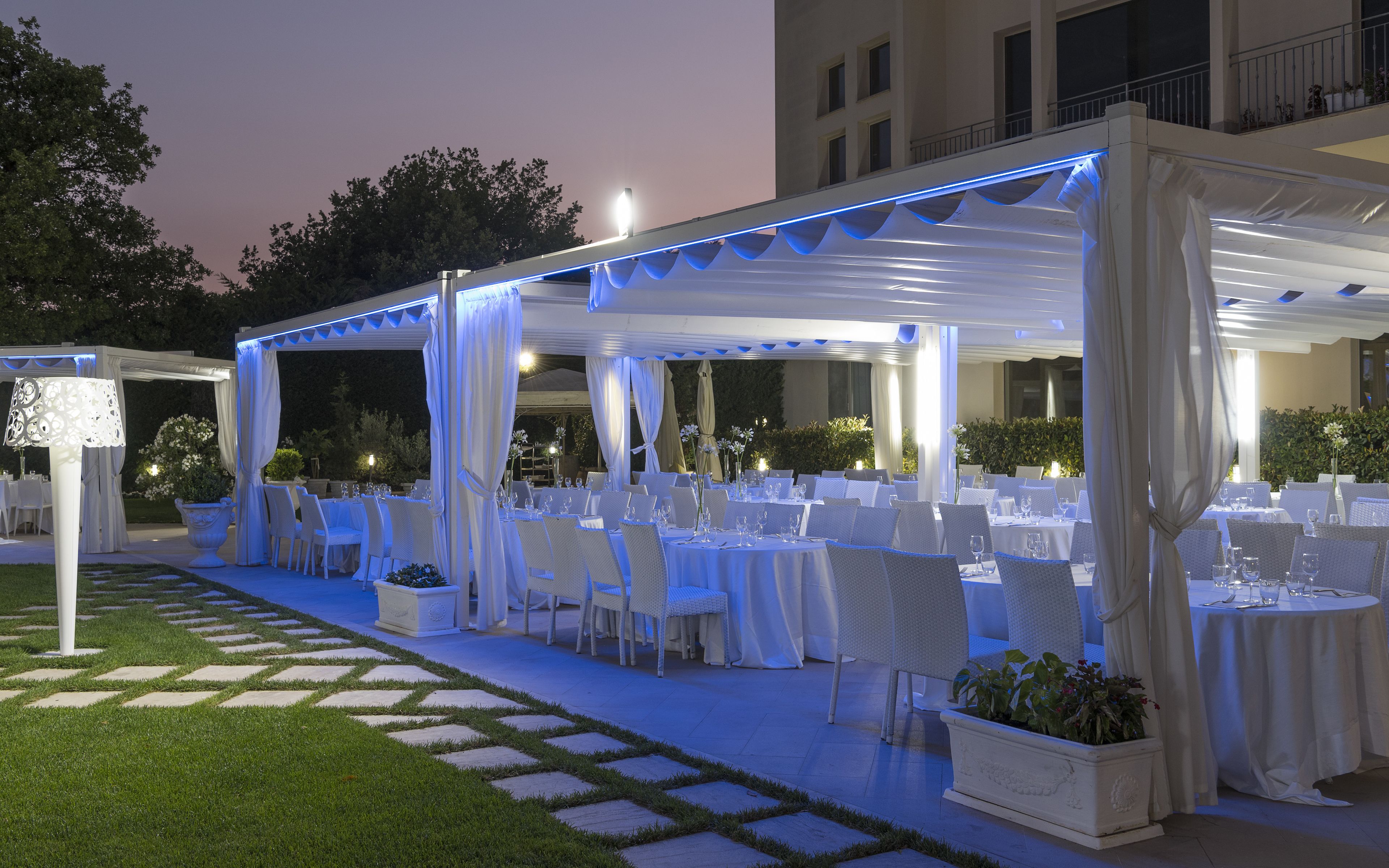 An outdoor dining area with white tables and chairs, covered by a white canopy with blue lighting, and surrounded by greenery.