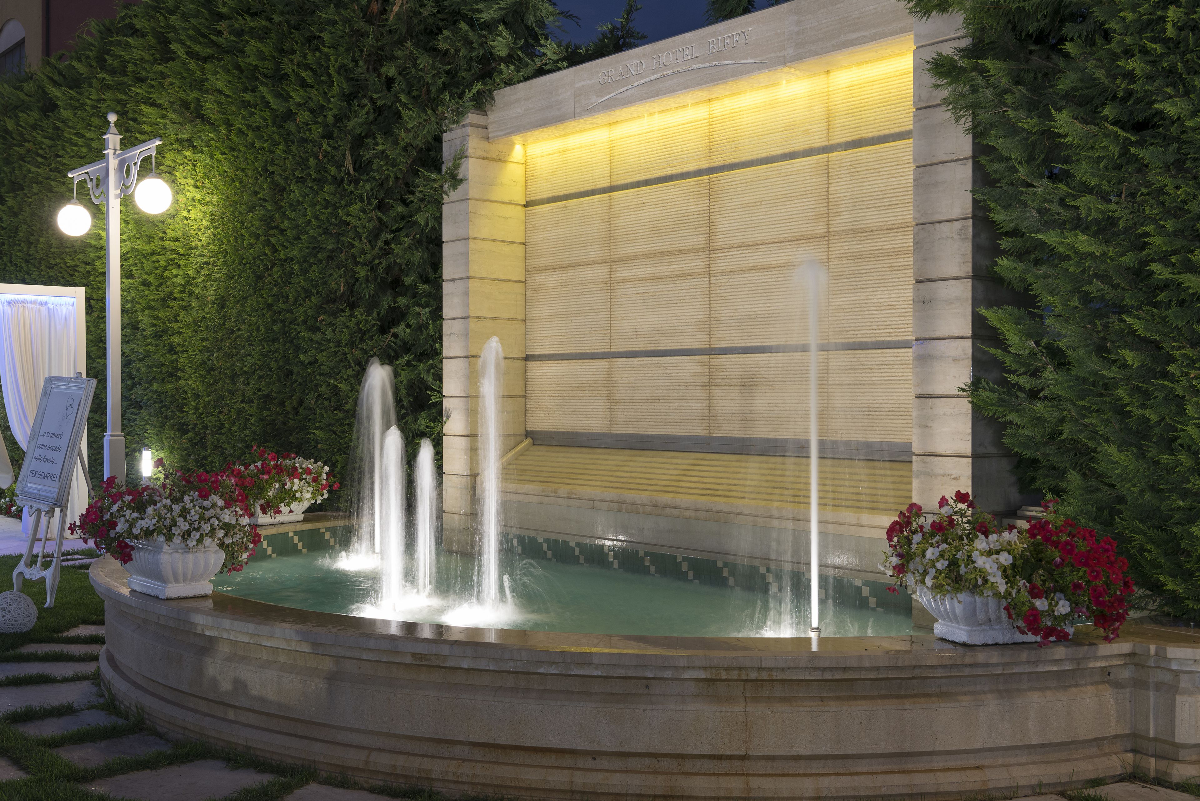A fountain with water jets, surrounded by a stone basin, and two flower pots with red and white flowers.