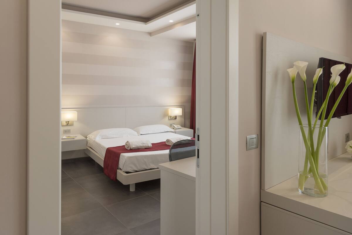 Ein modernes Hotelzimmer mit einem ordentlich bezogenen Bett, Nachttischlampen und einer Blumenvase auf dem Nachttisch.