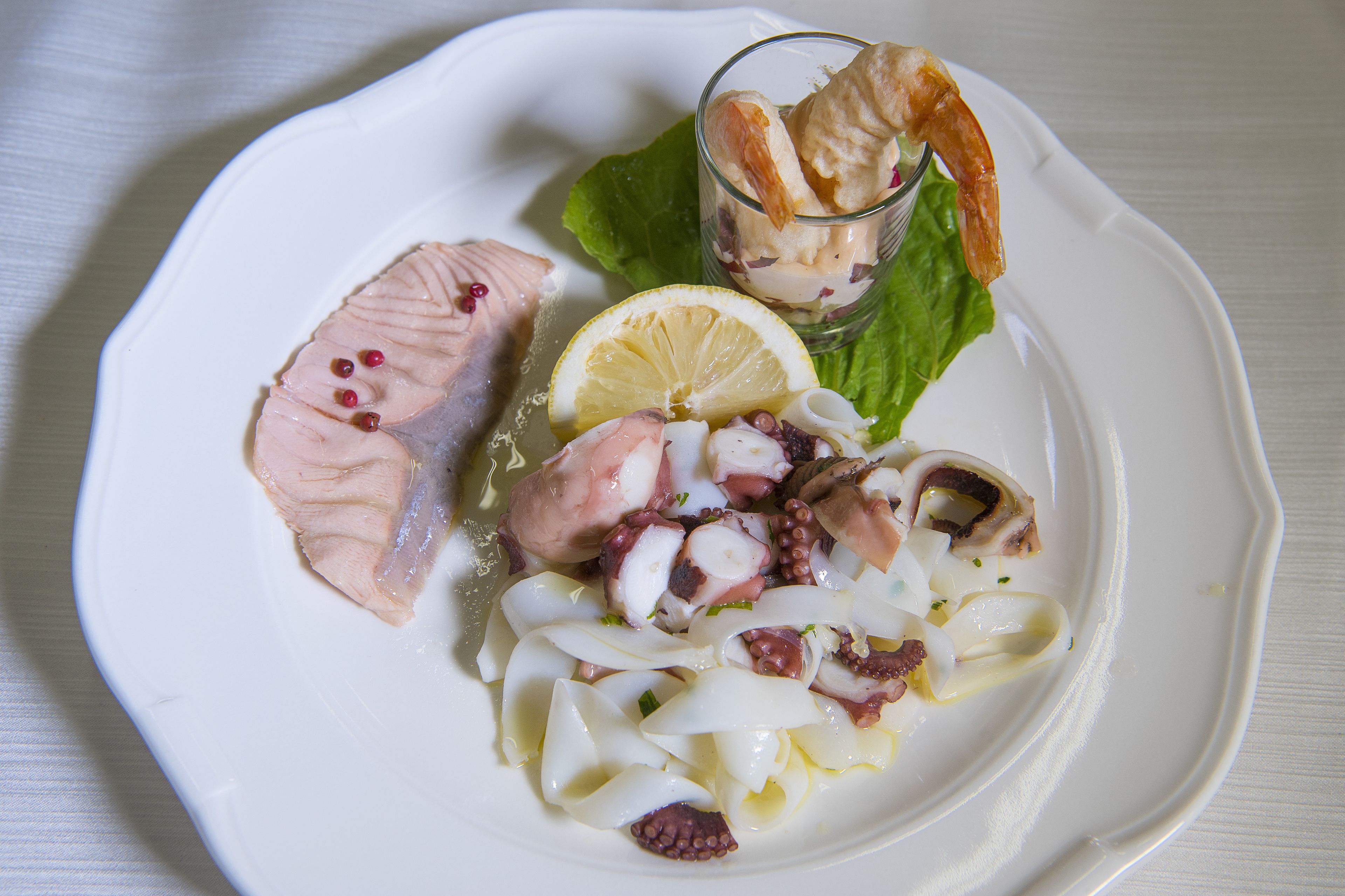 Un piatto con frutti di mare, tra cui polpo e gamberi, accompagnato da una fetta di limone e un bicchiere di salsa.