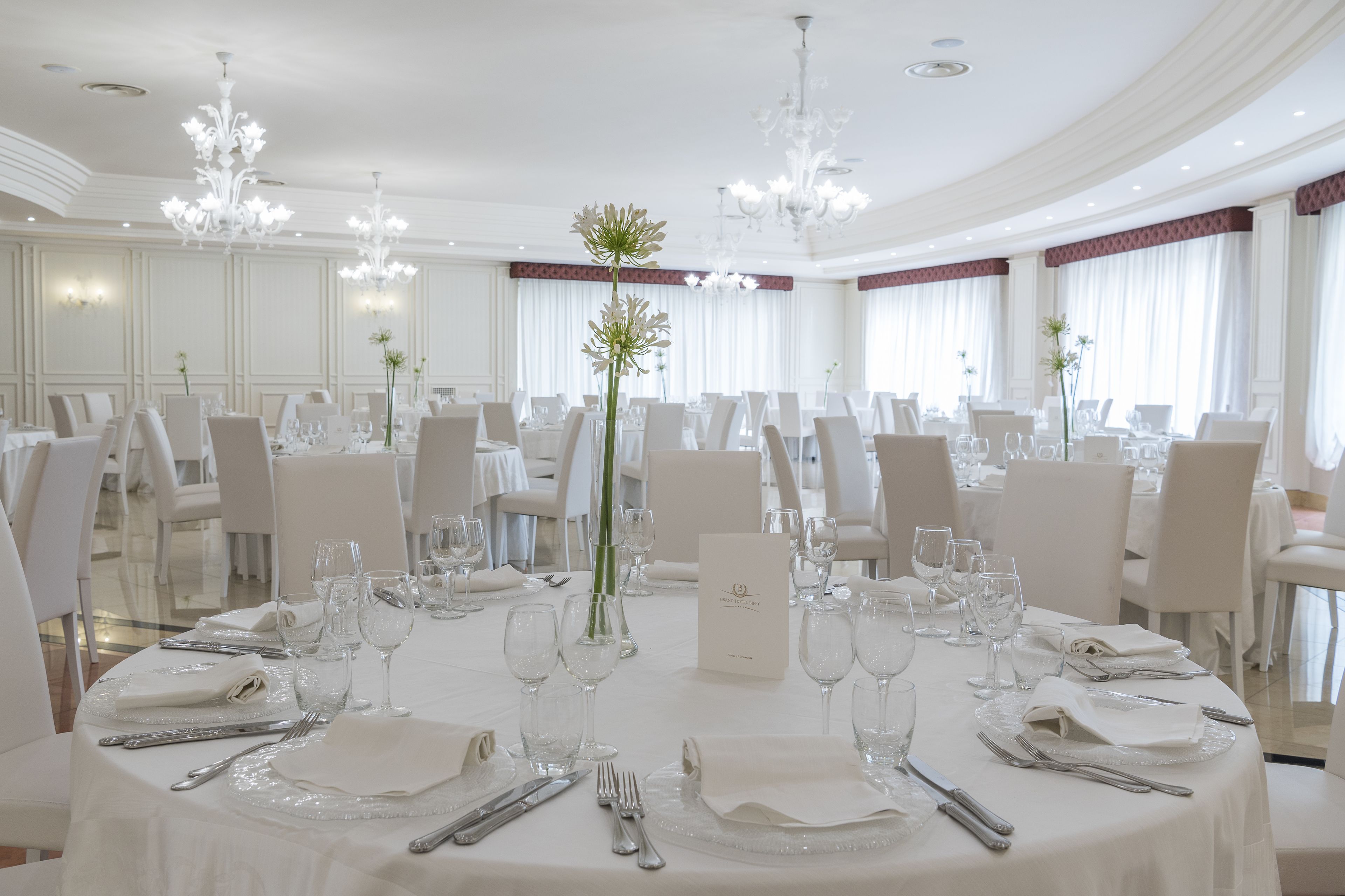 A formal dining room with round tables set for a meal, white chairs, and elegant chandeliers.