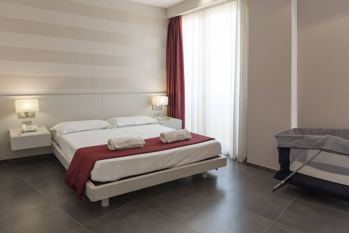 Ein modernes Hotelzimmer mit einem großen Bett, zwei Nachttischen mit Lampen, einem Schreibtisch mit Stuhl und einem Fenster mit Vorhängen.