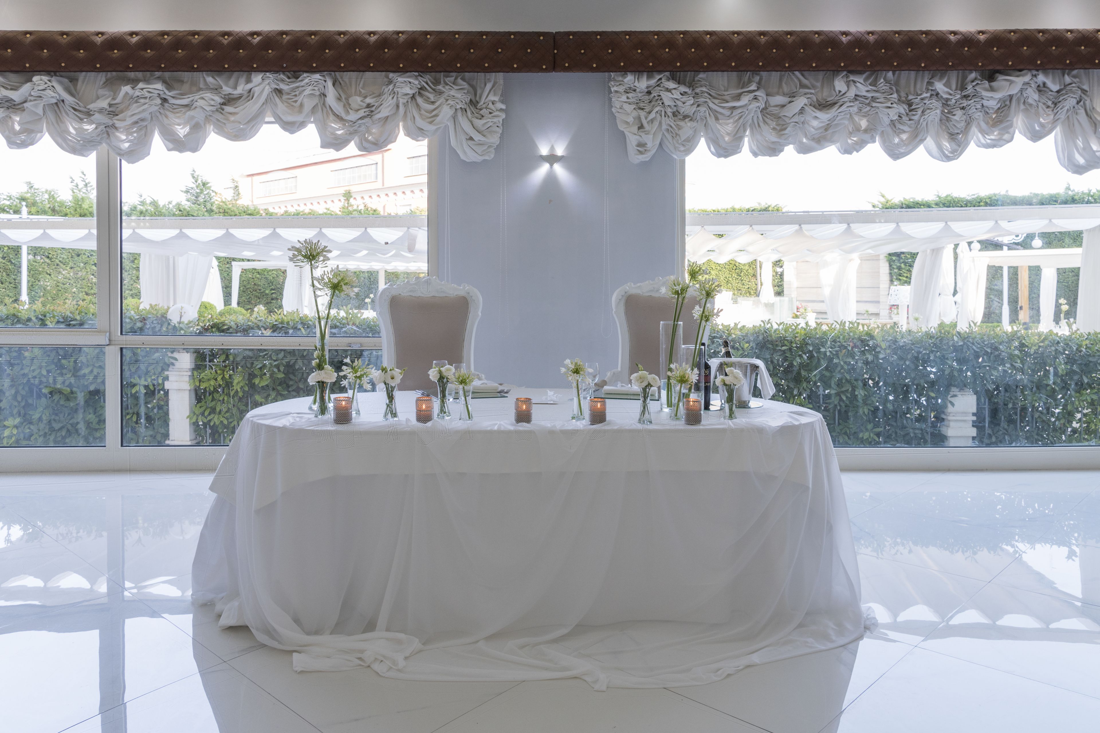 A round table with a white tablecloth, candles, and flowers, set against a large window with a view of greenery and buildings.