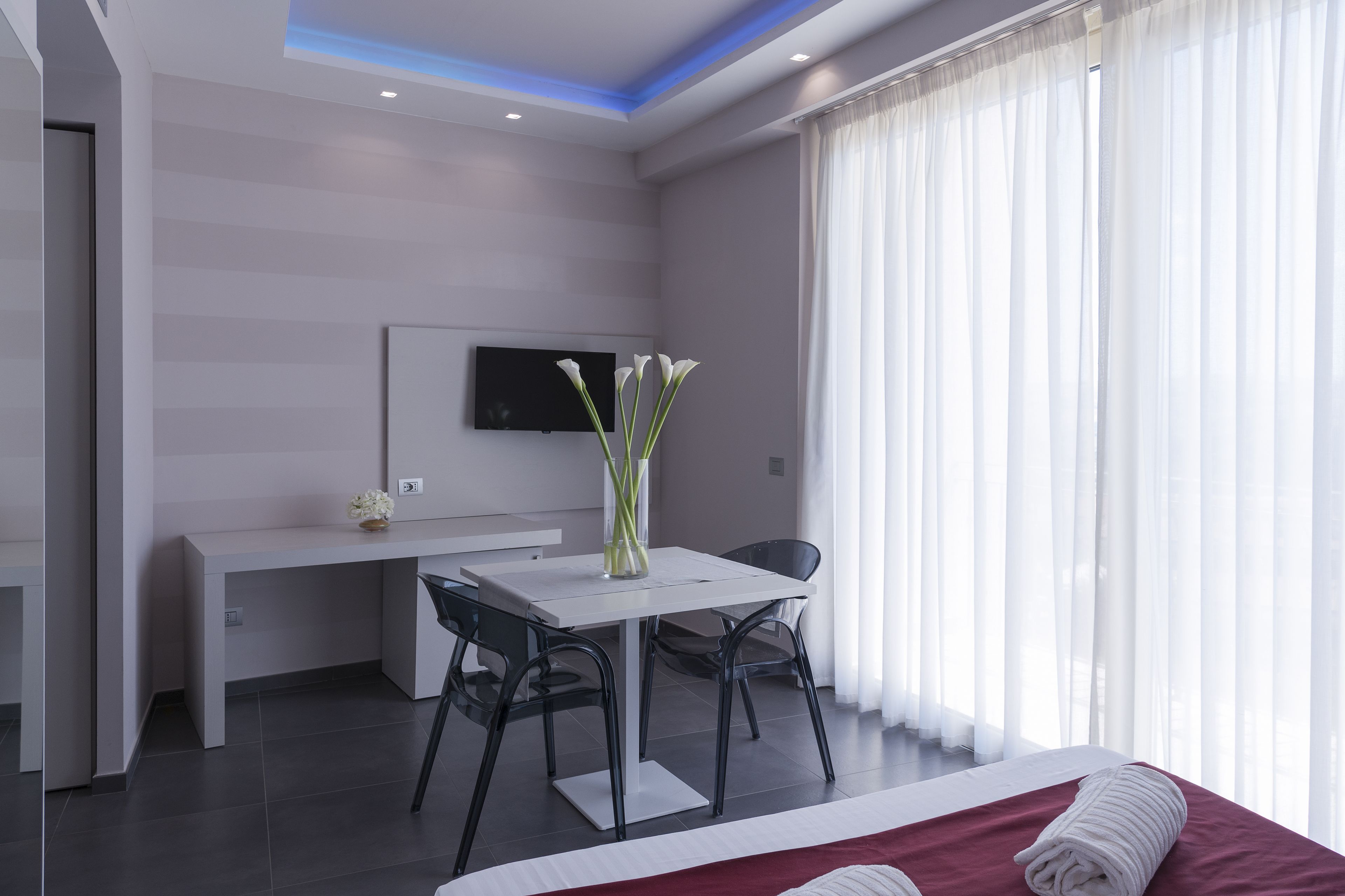 A modern hotel room with a bed, a desk, a chair, a flat-screen TV, and a vase with flowers.