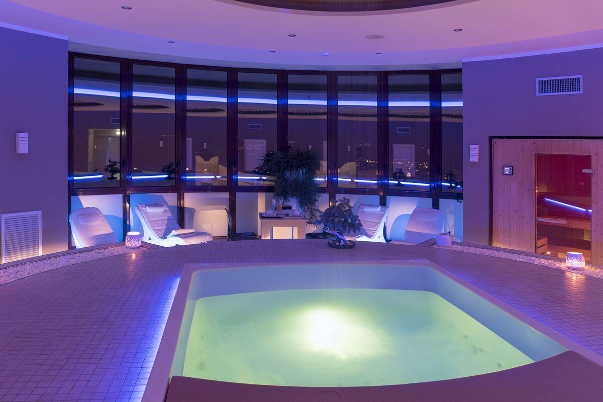 Ein Wellnessbereich mit großzügigem Whirlpool, Sitzgelegenheiten und Blick auf die Stadt durch große Fenster.