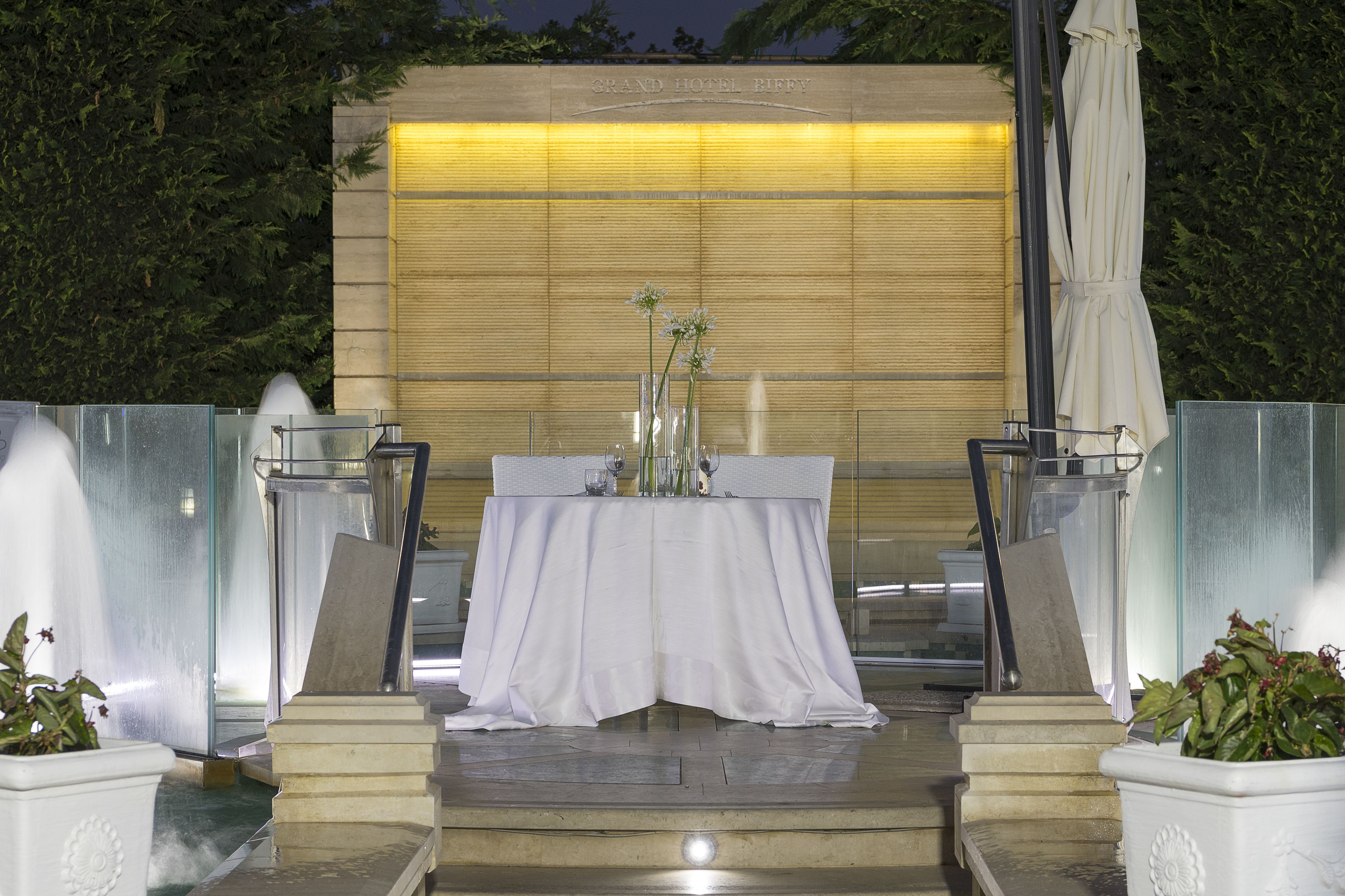 An outdoor dining area with a table set for two, surrounded by glass railings and potted plants.