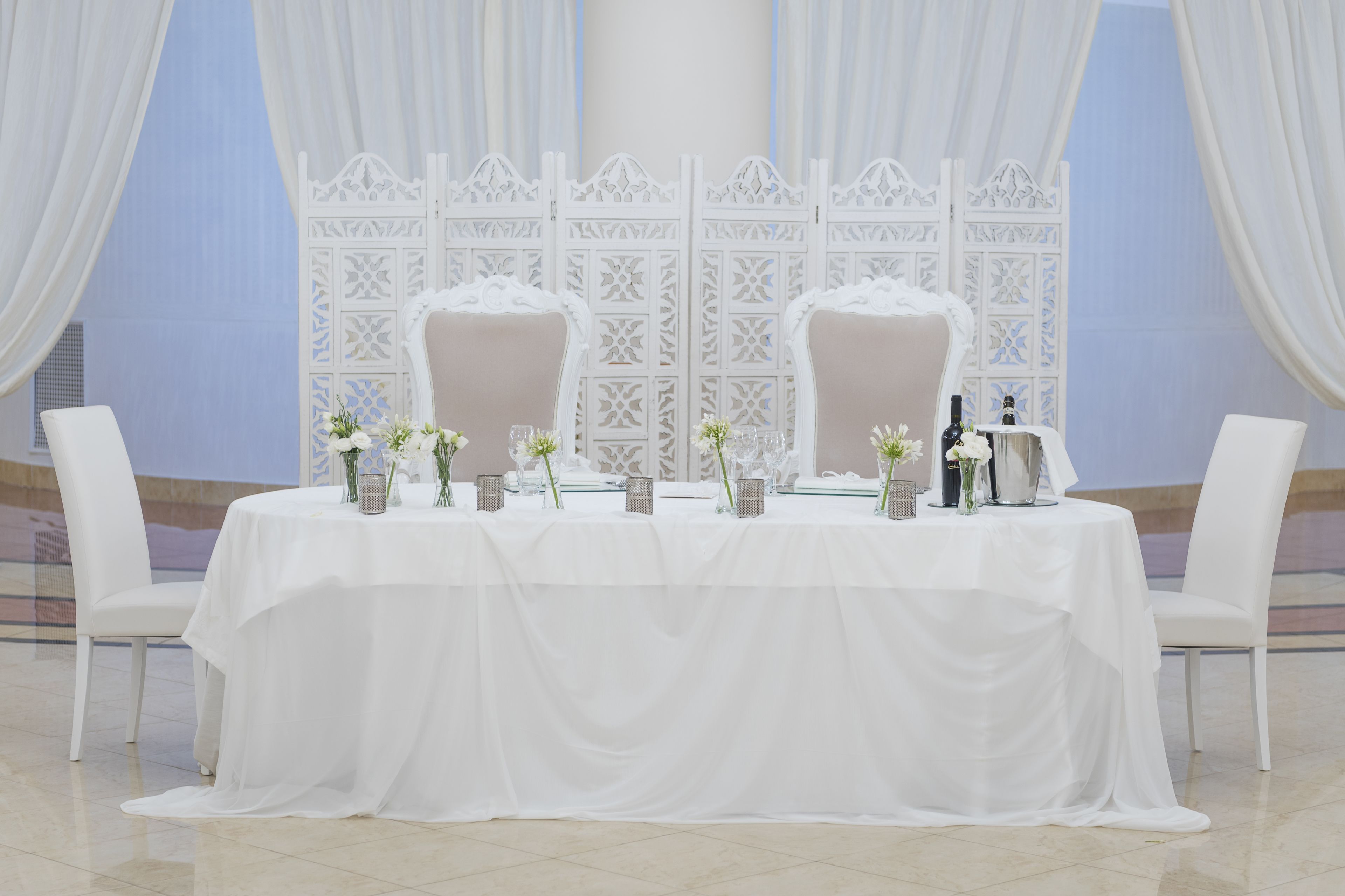 Mesa para dos con sillas blancas, mantel blanco y centros de mesa con flores.