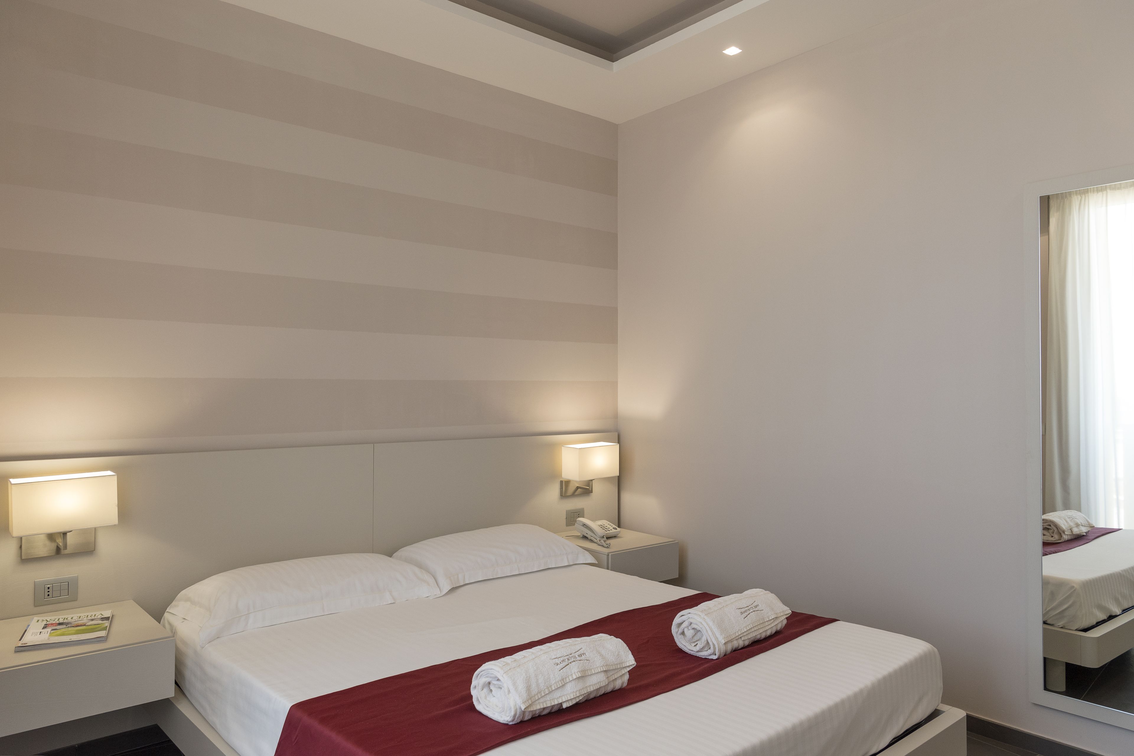 Une chambre d’hôtel avec un grand lit, deux tables de chevet avec des lampes, un miroir et une couverture rouge.