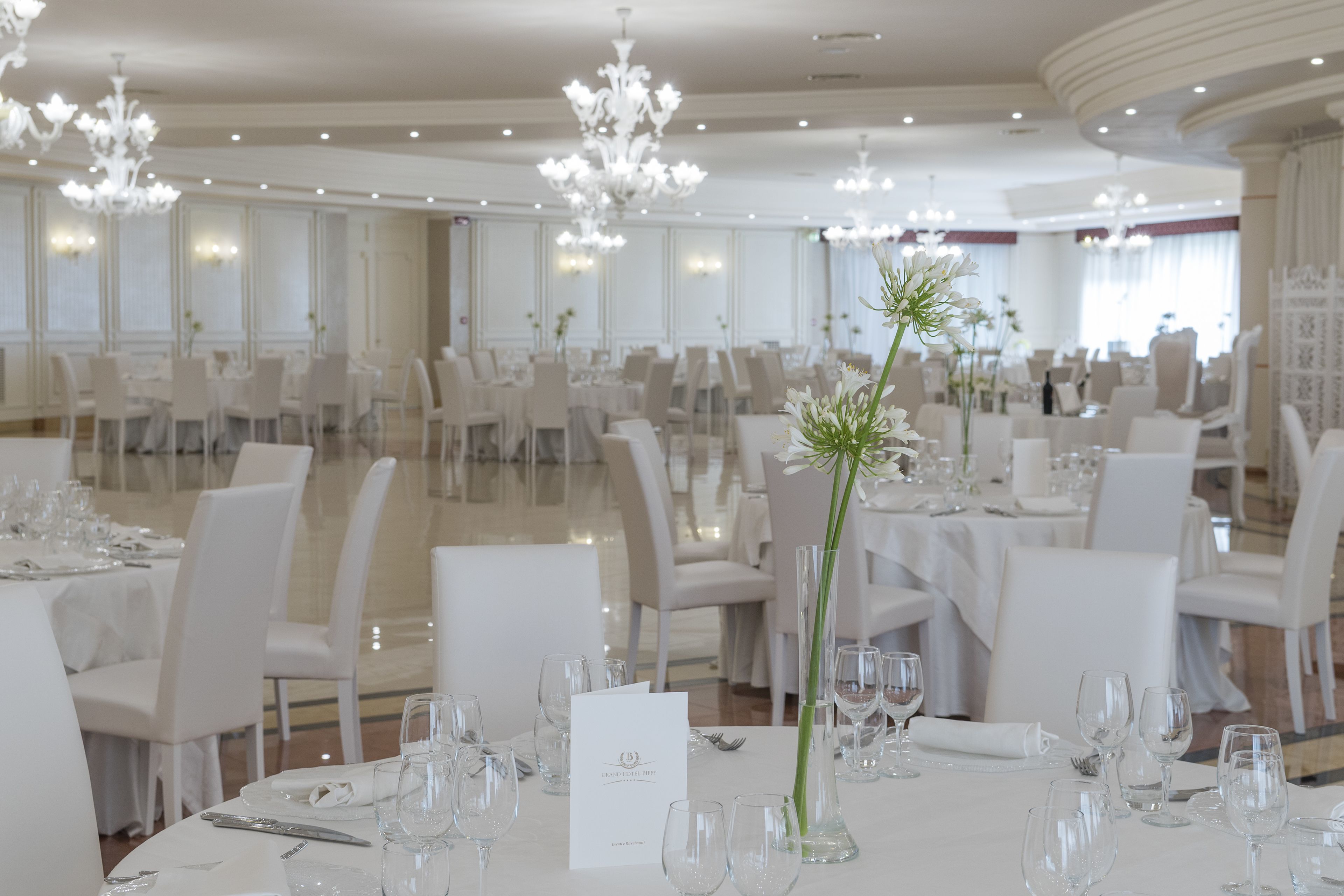 Une salle à manger formelle avec des tables, des chaises et des lustres élégants, le tout dans des tons blancs.