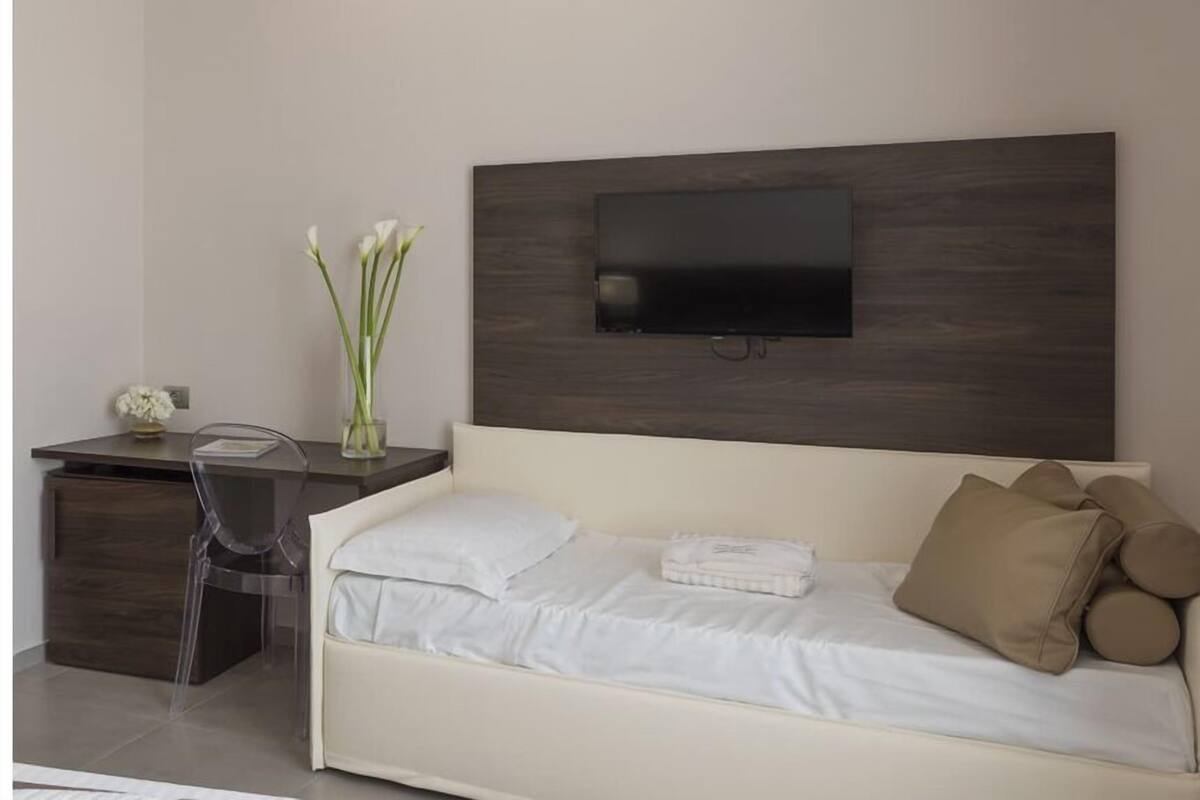 Ein modernes Hotelzimmer mit einem Bett, einem Schreibtisch, einem Stuhl, einem an der Wand montierten Fernseher und einer Vase mit Blumen.