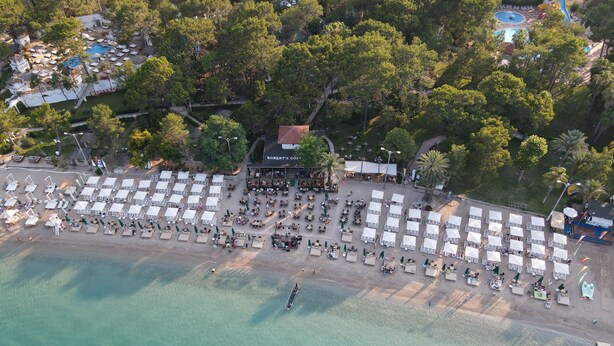 Vista aérea de un complejo turístico en la playa con filas de sillas de sol y sombrillas, una zona de piscina y un restaurante llamado "Robert's Coors".