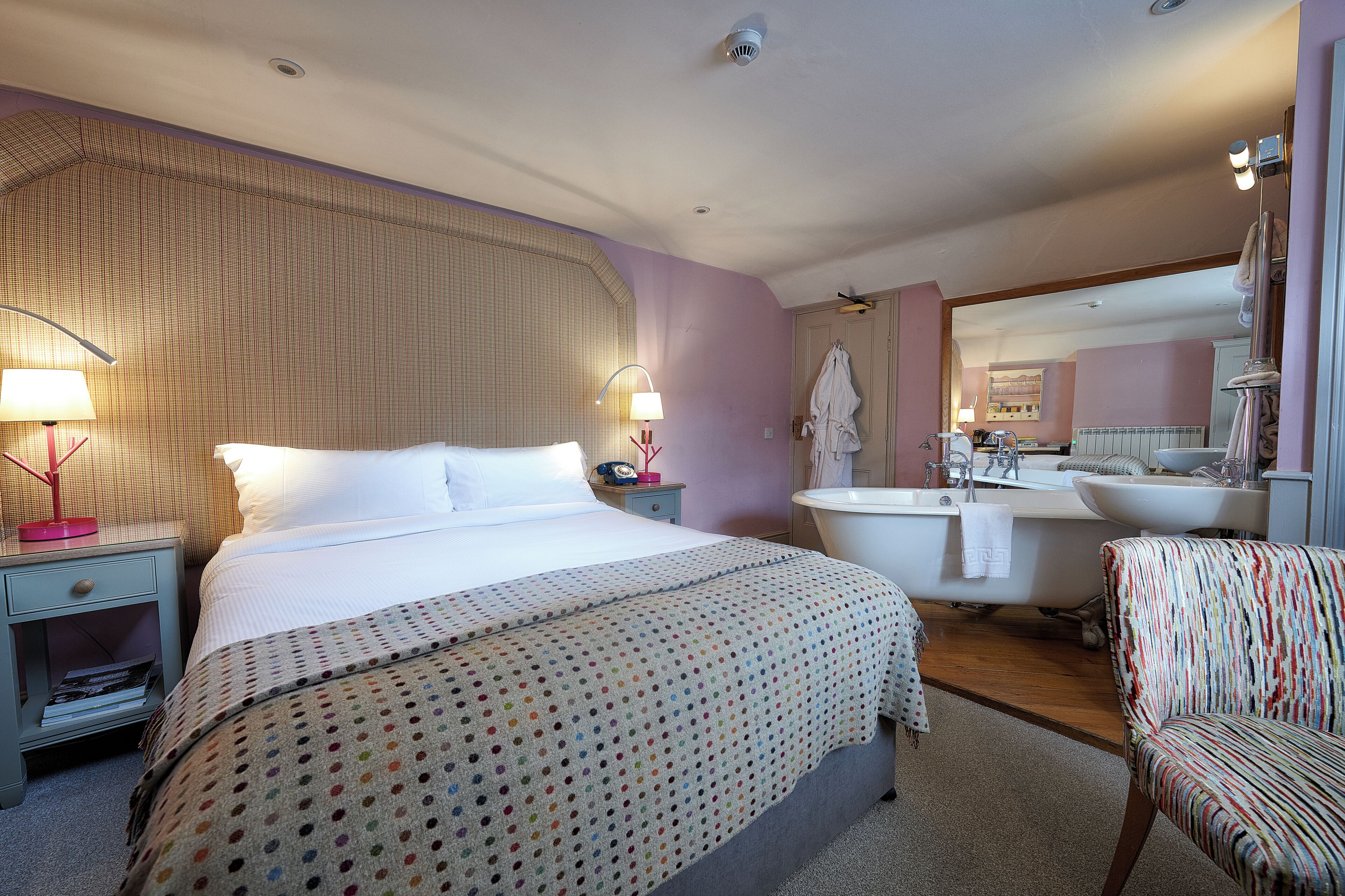 A hotel room with a bed, a nightstand, a lamp, a chair, a bathtub, and a mirror.