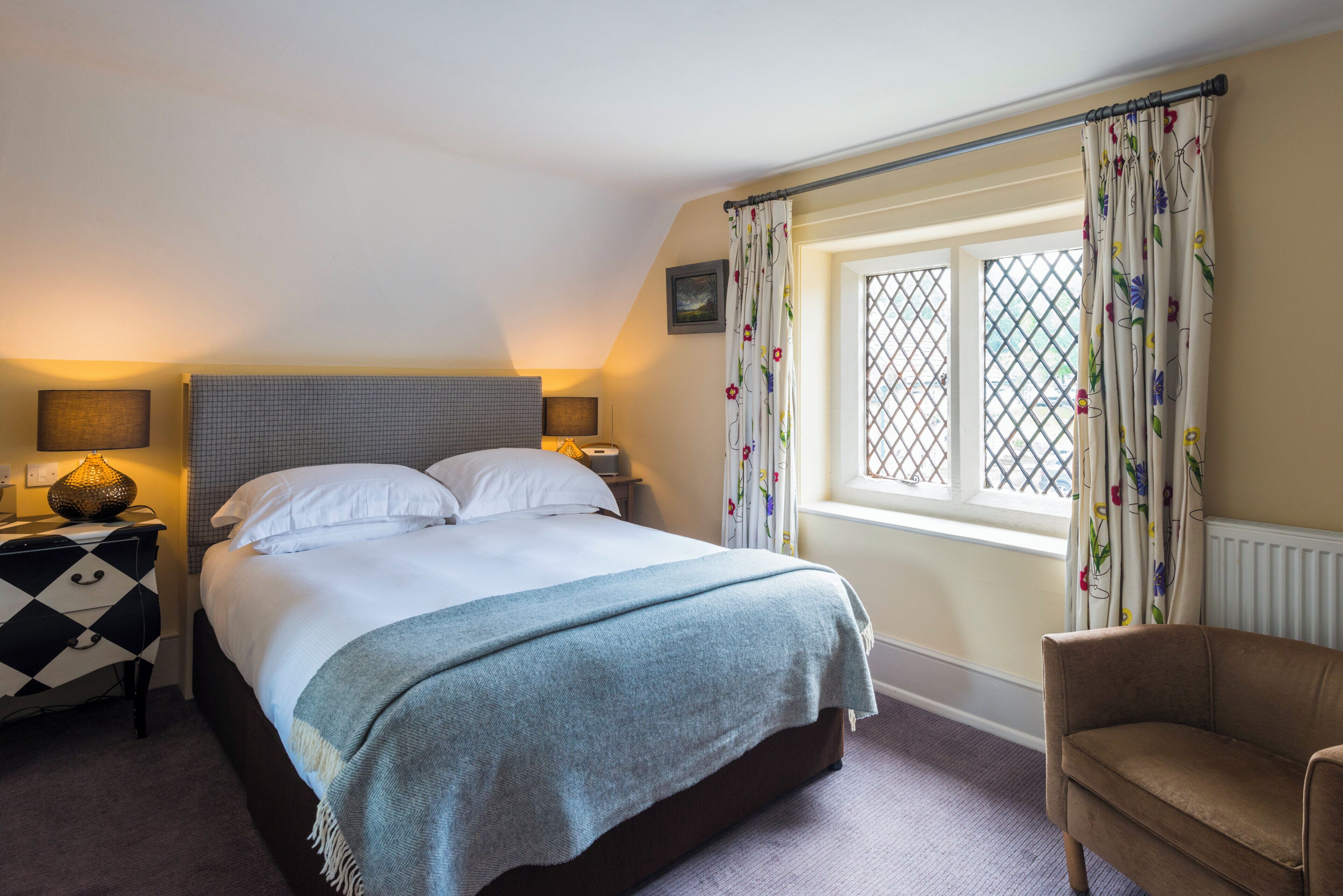 A neatly made bed with a grey blanket, a bedside table with a lamp, a chair, and a window with floral curtains.