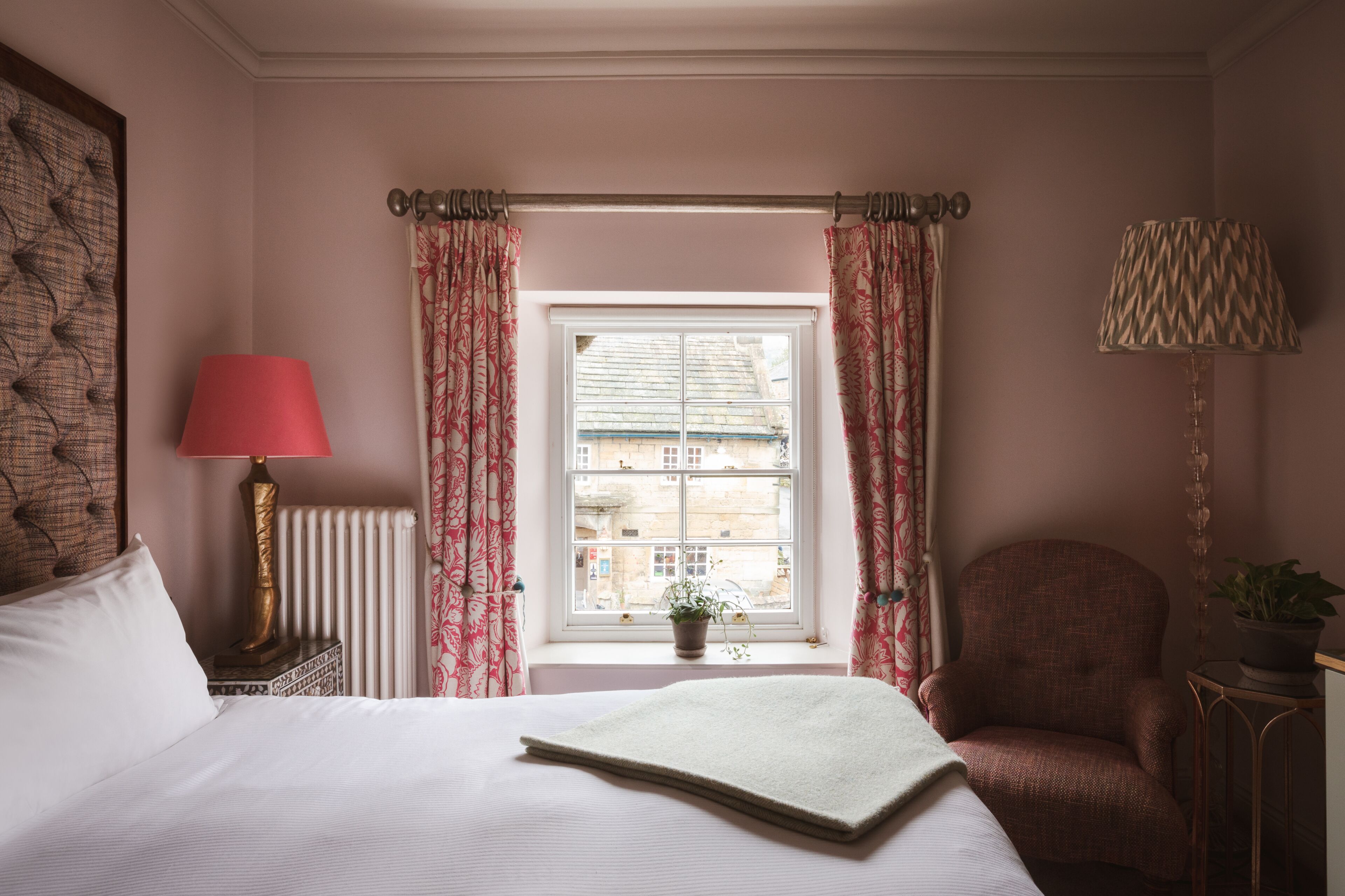 A bedroom with a bed, a nightstand with a lamp, a chair, a window with curtains, and a radiator.