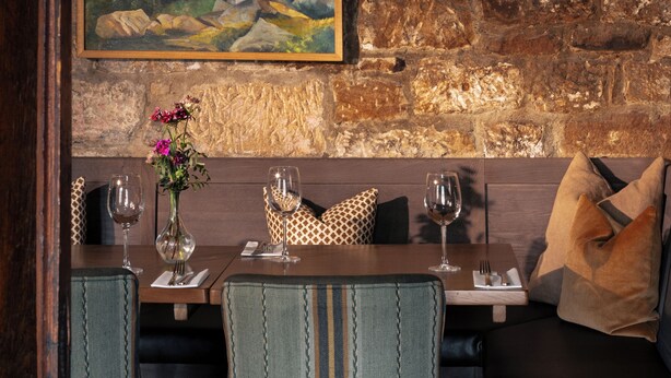A dining area with a rustic stone wall, a painting, and a table set for two with chairs. - The Beeley Inn  (Matlock)