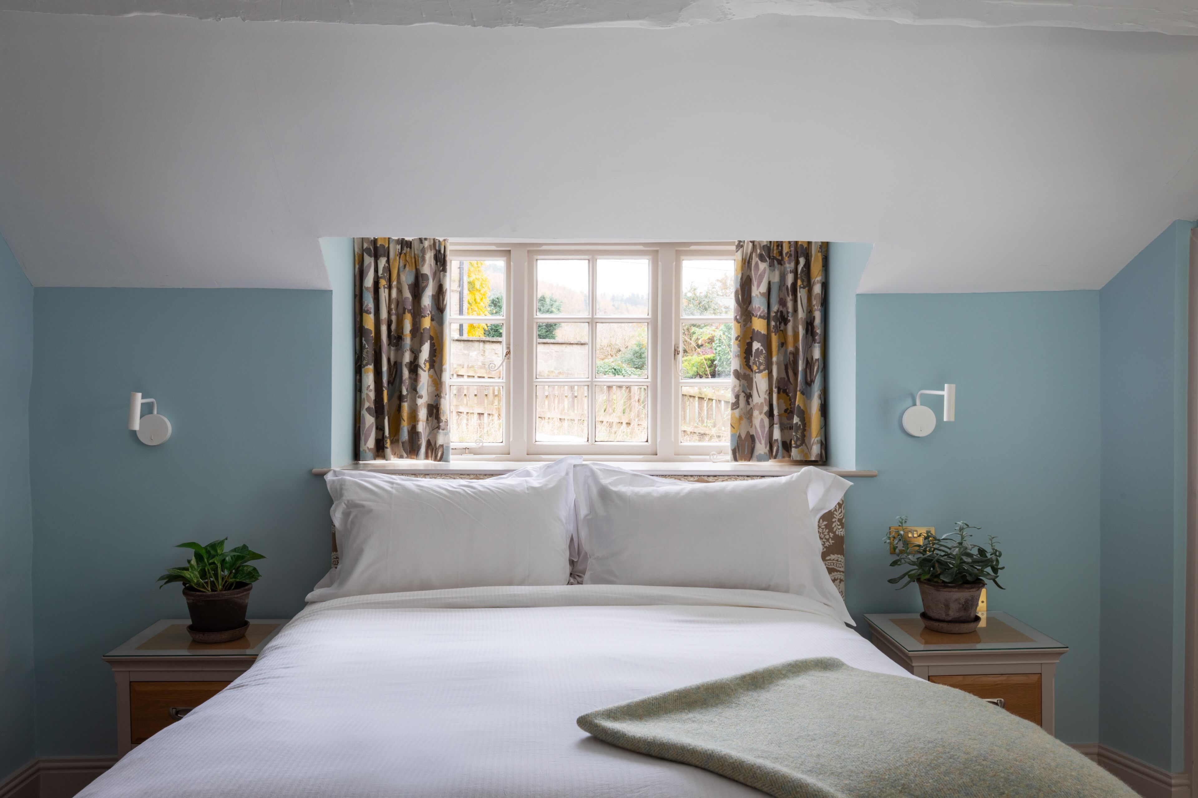 A bedroom with a bed, two bedside tables with potted plants, a window with curtains, and a wall-mounted light fixture.