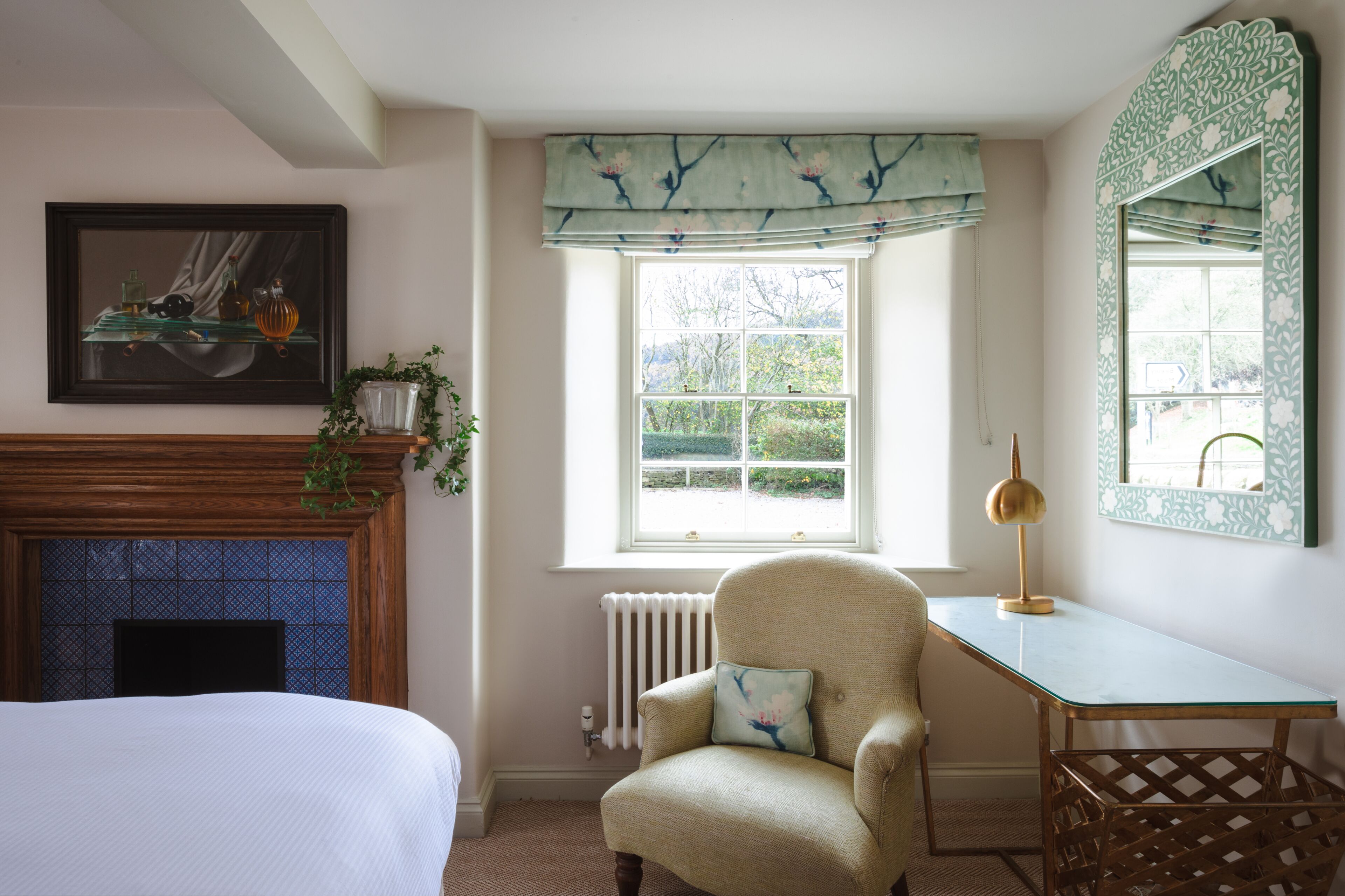 A bedroom with a bed, a chair, a desk, a fireplace, a window with a view, and a mirror.