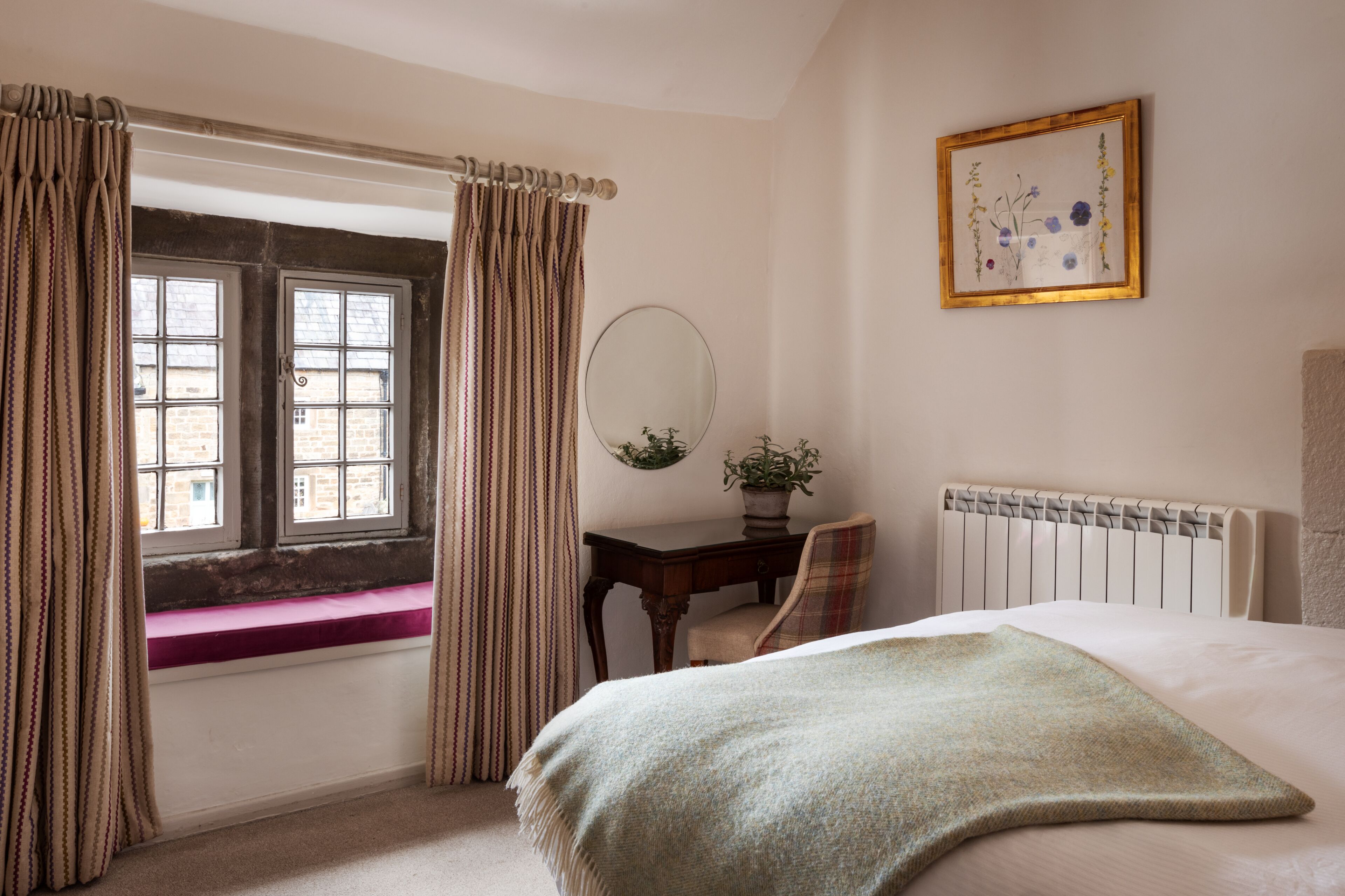A bedroom with a window, a bedside table with a mirror, a radiator, and a bed with a green blanket.