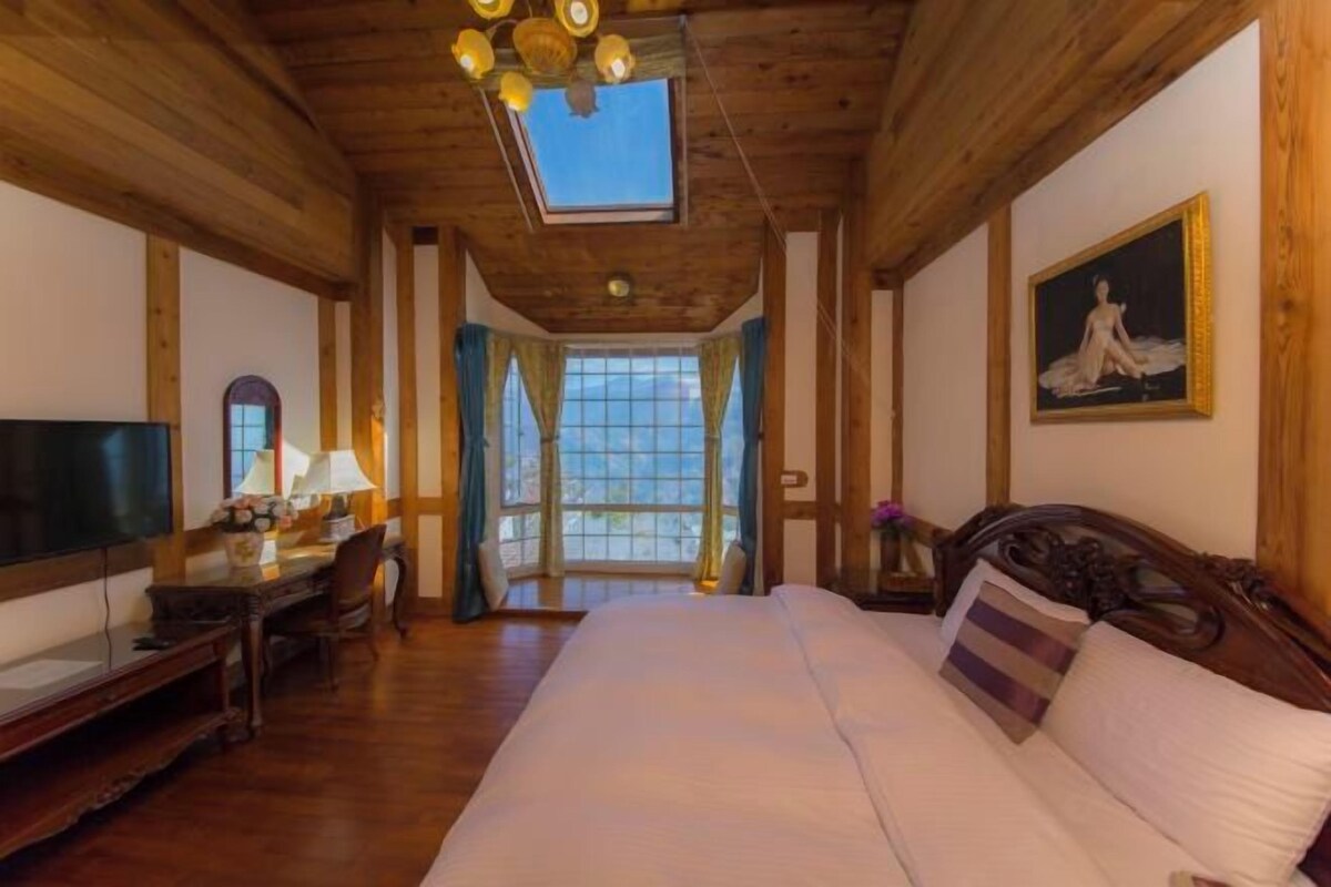 Una habitación con una cama grande, techo de madera, un escritorio con lámpara, televisor y un cuadro en la pared.