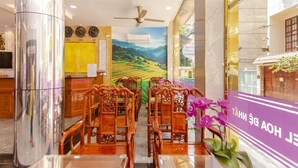 A hotel lobby with a mural of a mountainous landscape, wooden chairs and tables, a reception desk, and a purple sign with text. - Hoa De Nhat Hotel (Ho Chi Minh City)