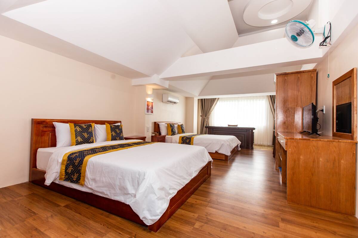 Habitación de hotel con dos camas, suelo de madera, ventilador de techo, televisor y escritorio.