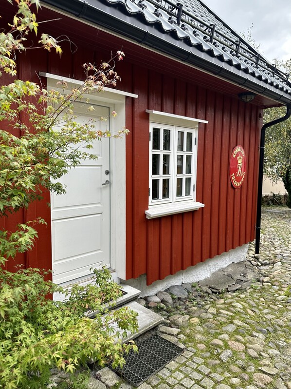 A red building with a white door and window, a stone pathway, and a metal grate. - Toldgaarden Gjestegaard (Larvik)