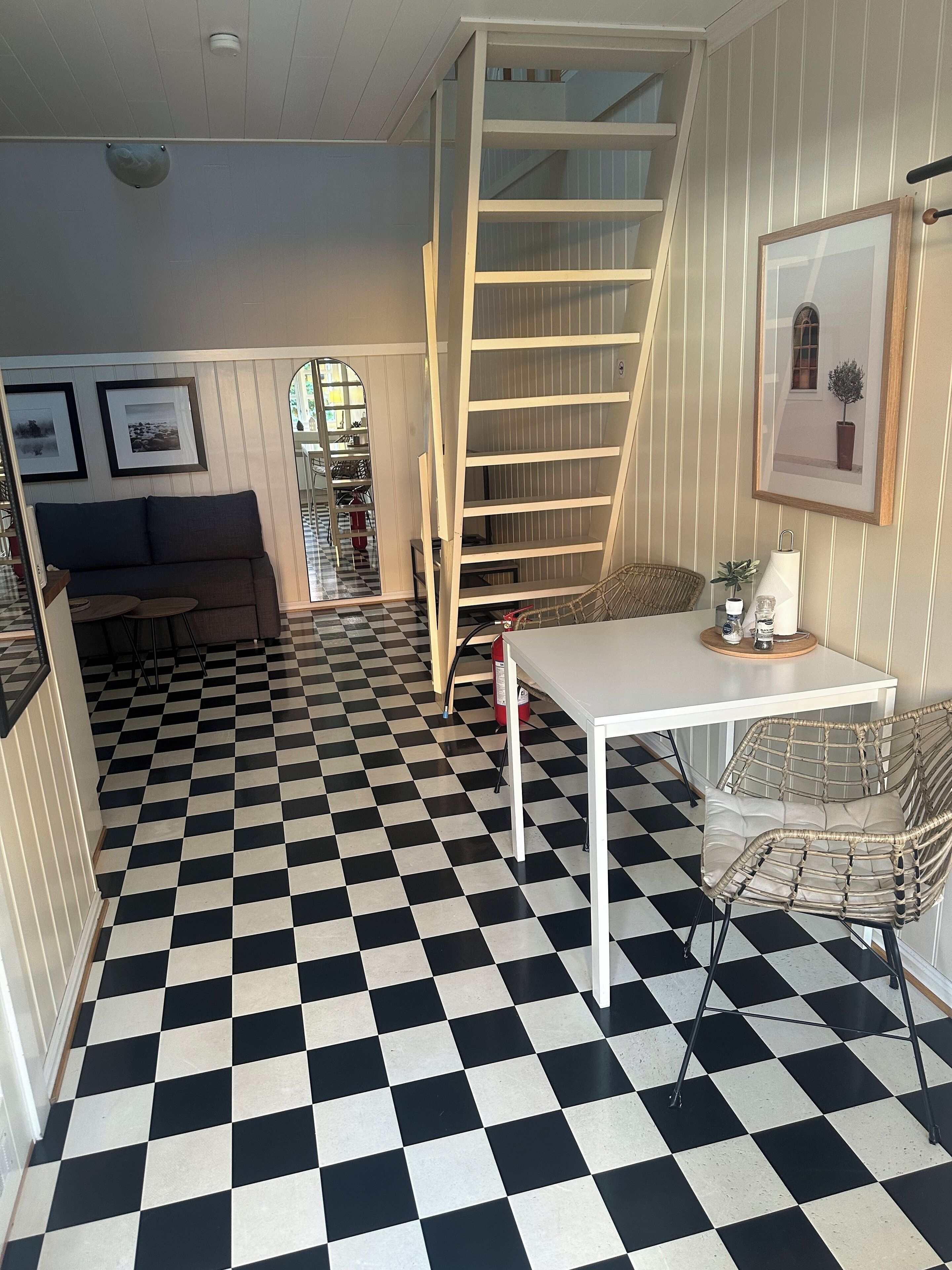 A room with a checkered floor, a white table, a wicker chair, a sofa, a staircase, and framed pictures on the wall.