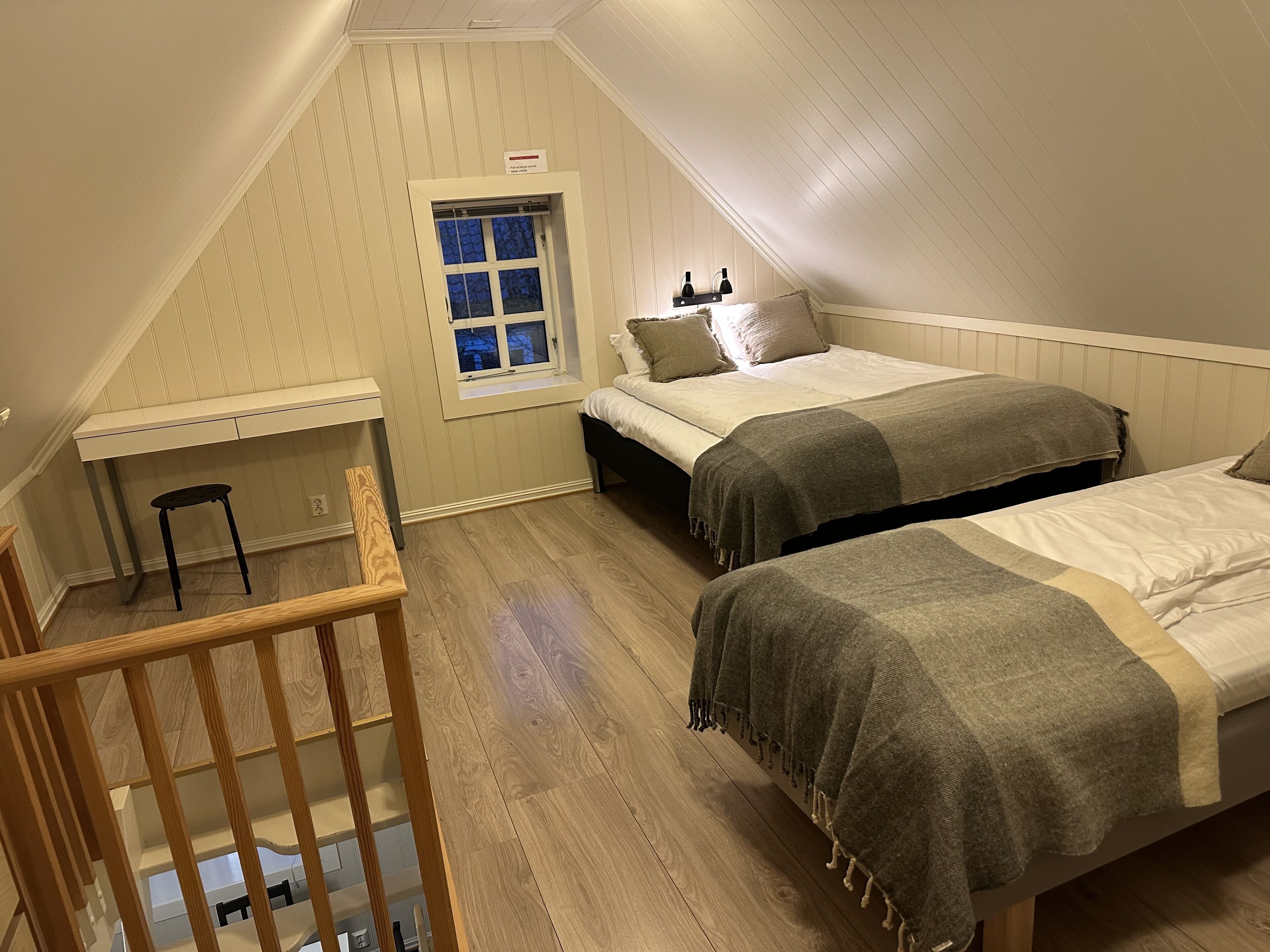 A dormitory room with two beds, a desk, a chair, and a window.