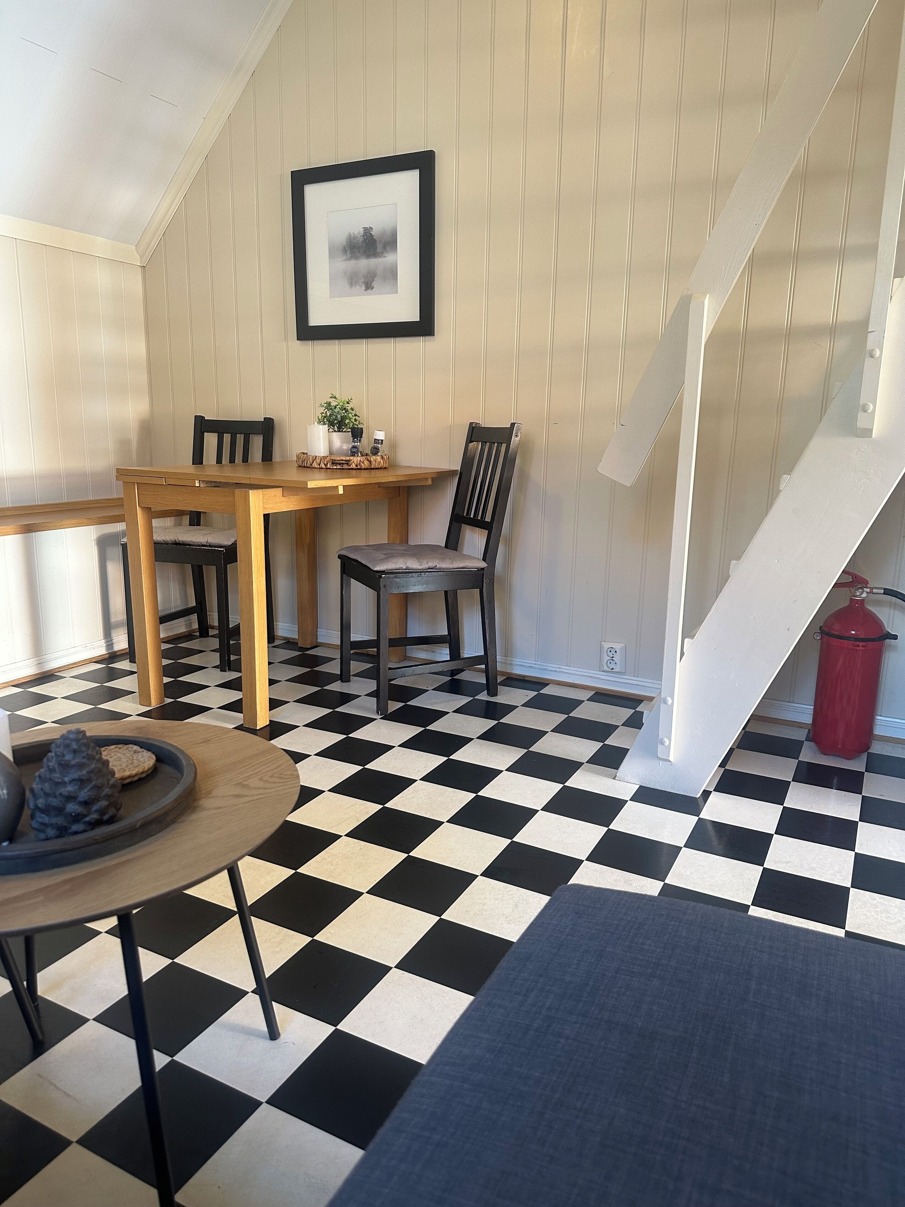 Een eetgedeelte met een zwart-wit betegelde vloer, een houten tafel, twee stoelen, een lijst met een foto aan de muur en een trap.