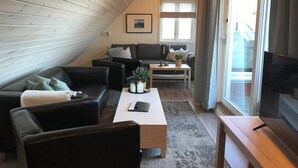 A cozy loft-style living room with a sloped ceiling, a large window with curtains, a flat-screen TV, and a fireplace. - Toldgaarden Gjestegaard (Larvik)