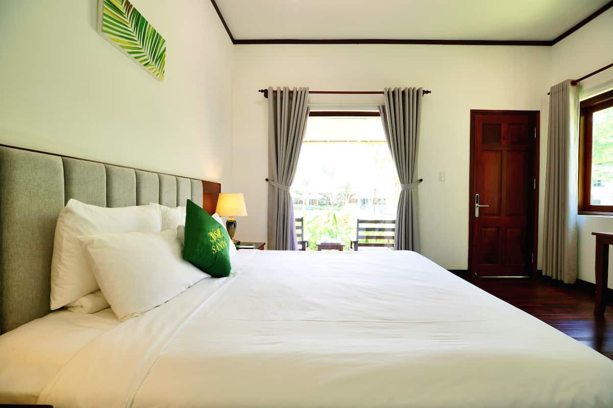 A neatly made bed with white linens and a green pillow, a wooden headboard, a window with curtains, and a view of greenery outside.
