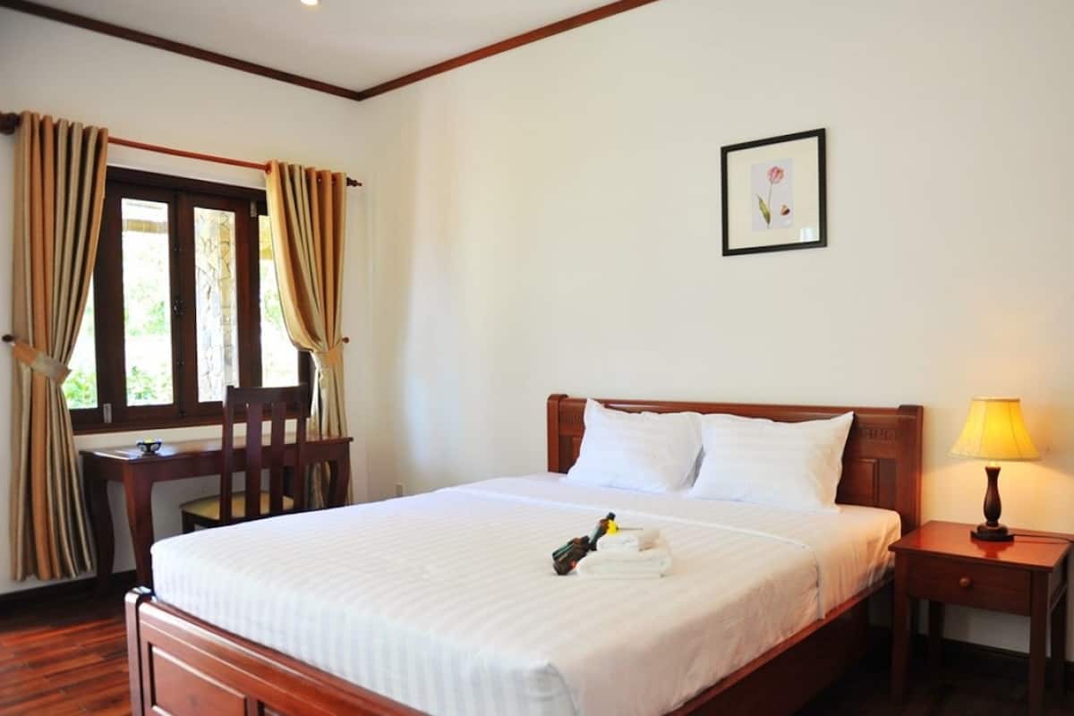 A hotel room with a bed, a desk, a chair, a window with curtains, a bedside lamp, and a framed picture on the wall.