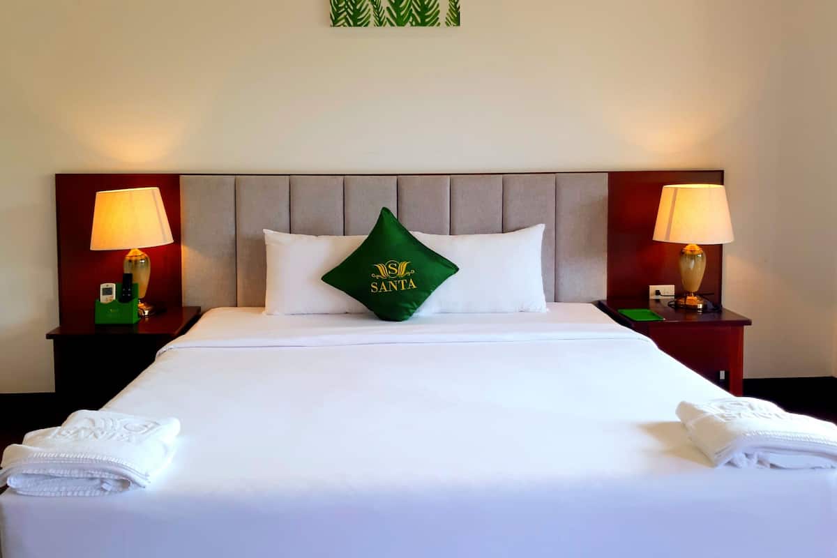 A hotel room with a large bed, two nightstands, a green pillow with 'SANTA' text, and two lamps.