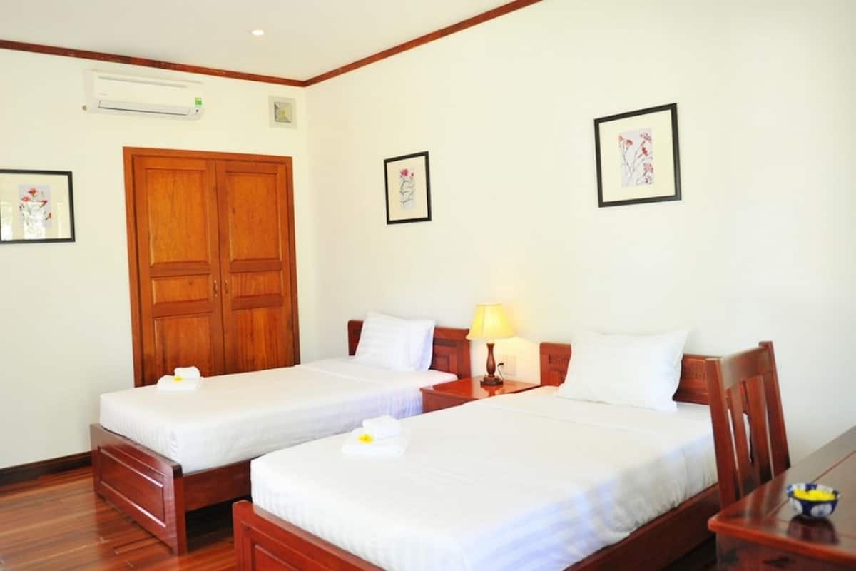 A hotel room with two single beds, wooden headboards, a wooden wardrobe, a bedside table with a lamp, and framed artwork on the walls.
