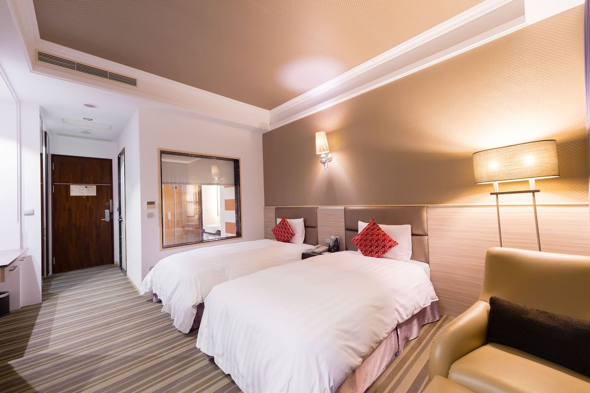 Habitación de hotel con dos camas, un área de descanso con una silla, un espejo, una lámpara de pared y una puerta que da a otra habitación.