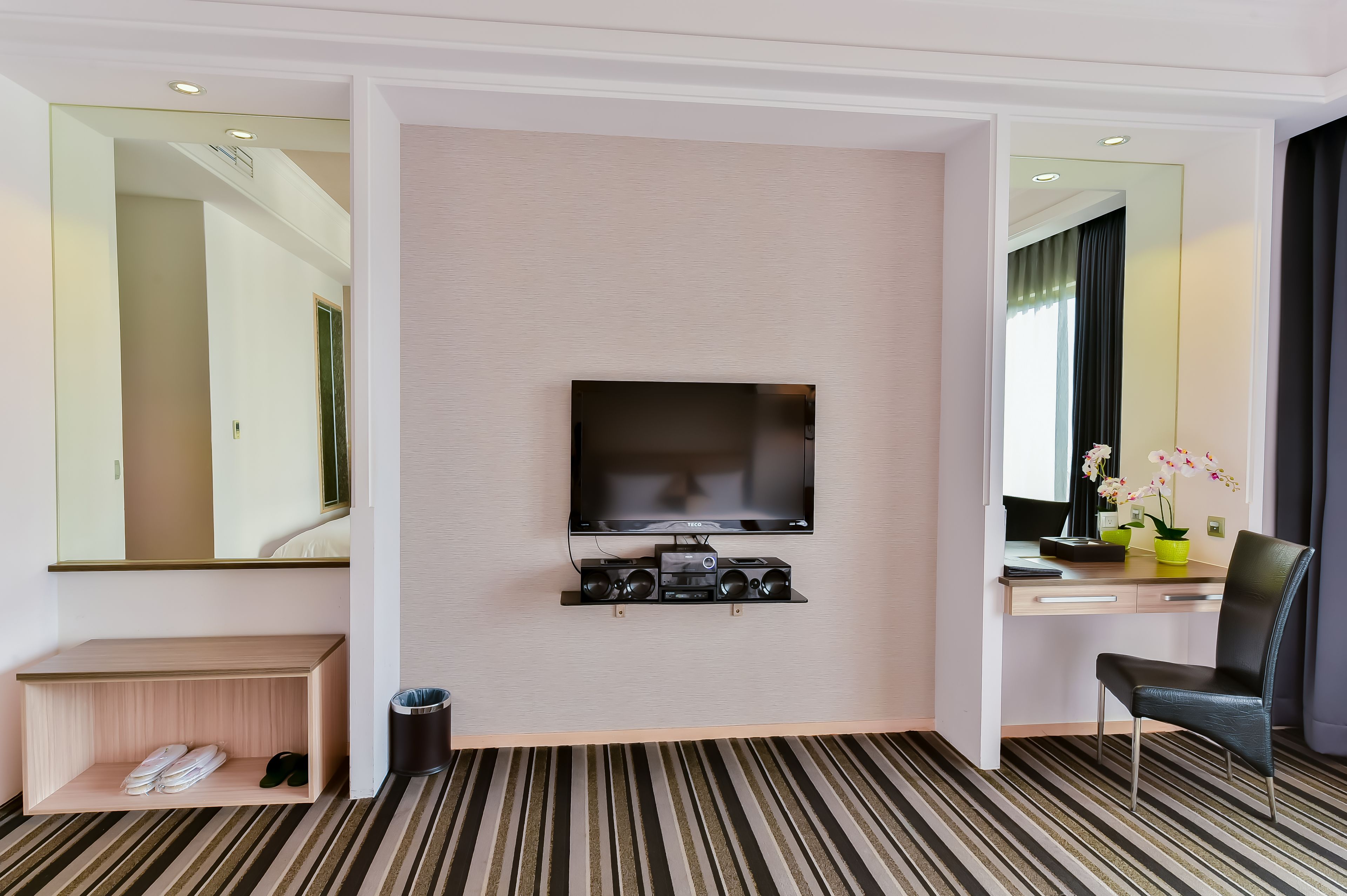 Habitación de hotel con televisor de pantalla plana montado en la pared, un escritorio con silla, un estante con zapatos y un cubo de basura.